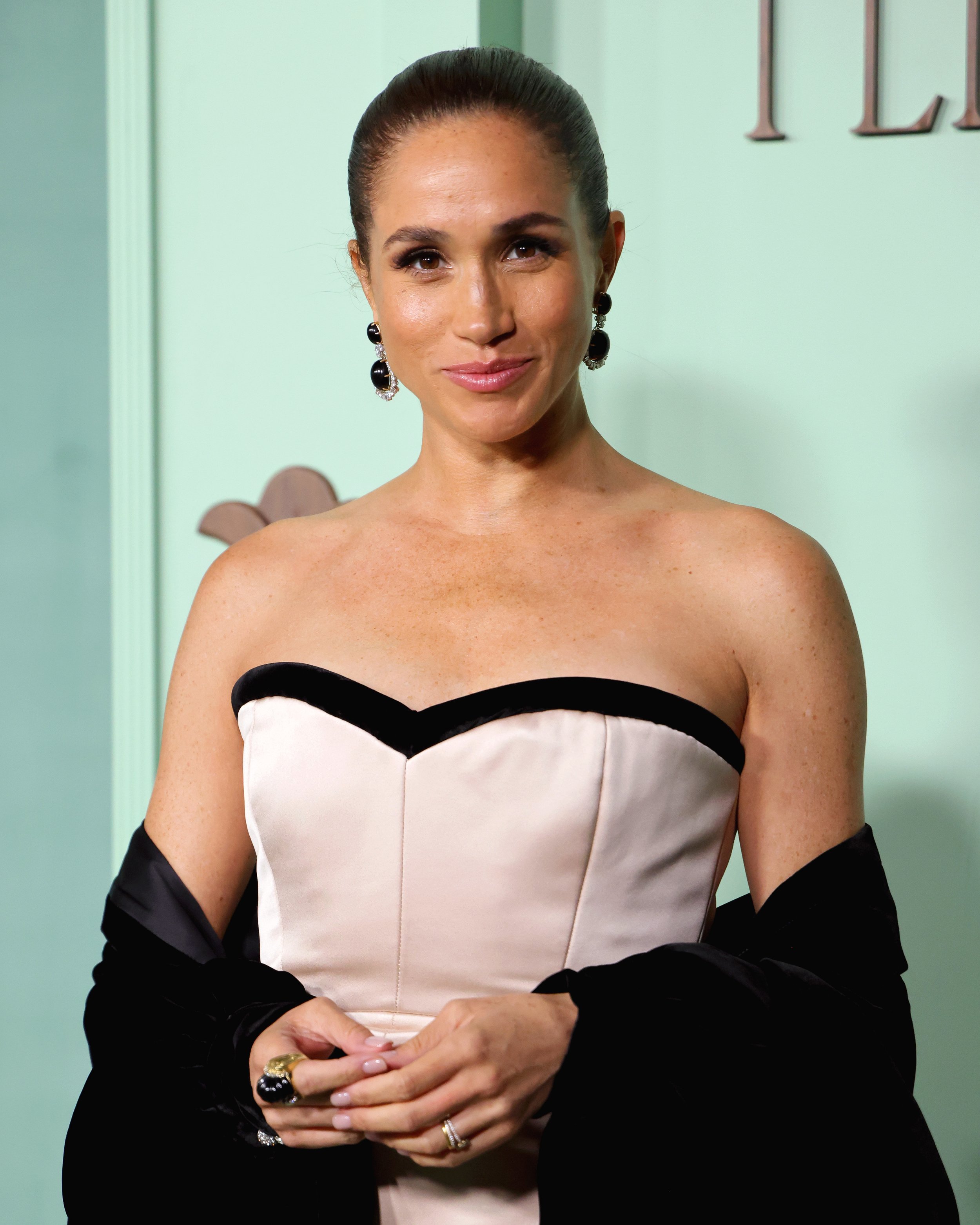 A woman wearing a strapless beige dress with black accents, black earrings, and rings, standing indoors against a pale green wall.