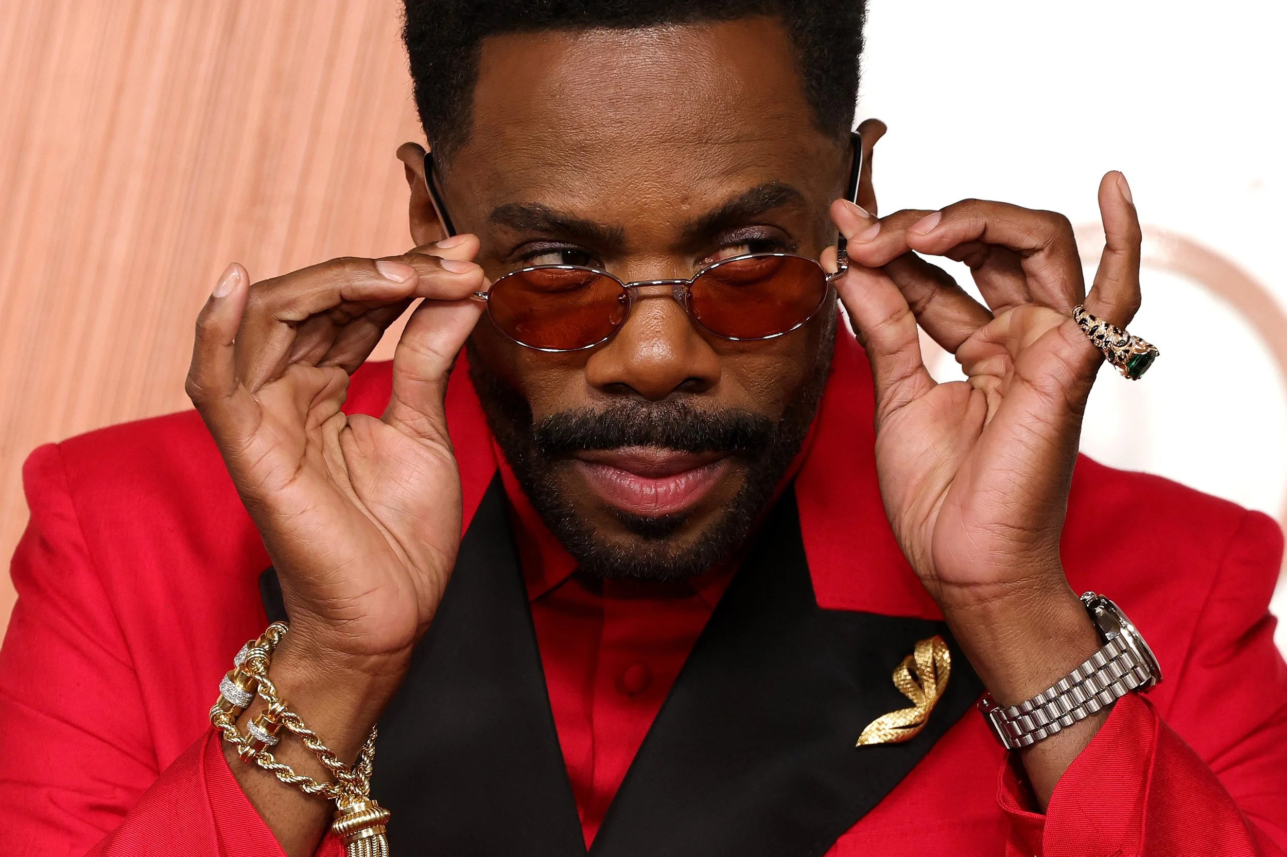 A man with a well groomed beard wearing a red suit, black shirt, and accessories, is pulling down tinted glasses to reveal his eyes, looking confident.