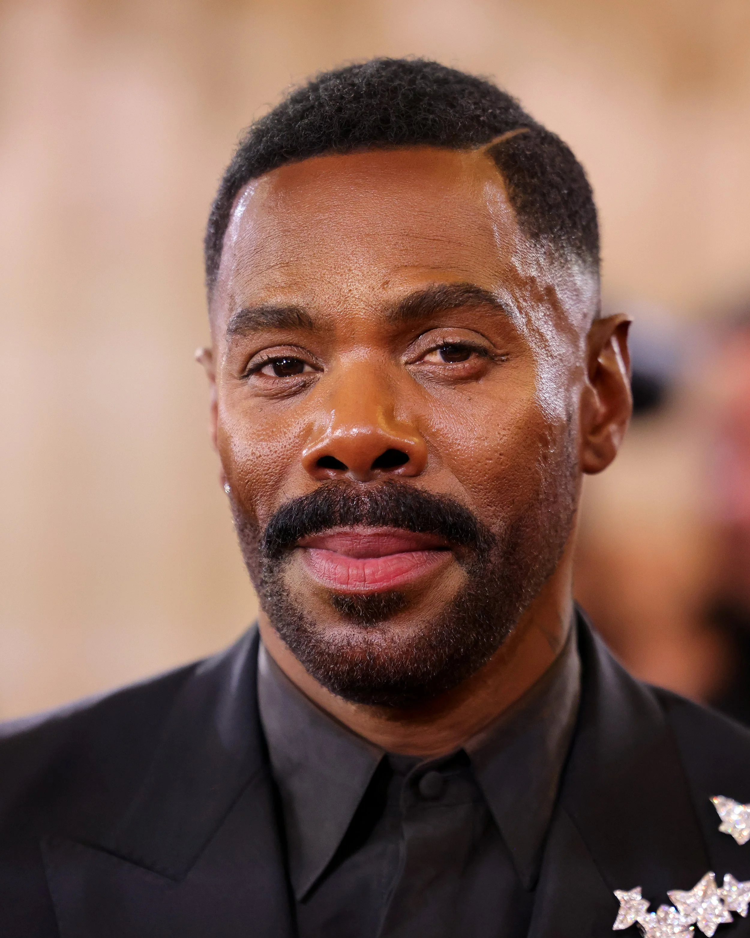 Close-up of a man with dark skin, short hair, mustache, and beard, wearing a black suit with a star-shaped badge.