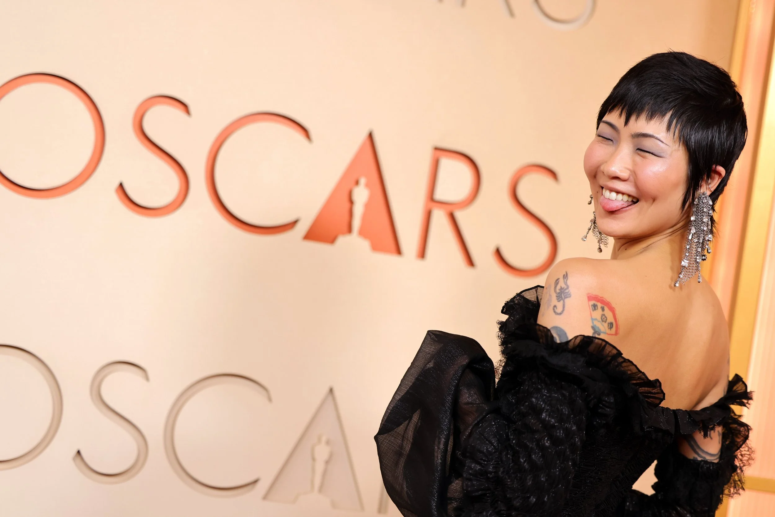 A woman with short black hair and dangling earrings smiling and sticking out her tongue at an Oscars event, with tattoos visible on her shoulder and upper arm.