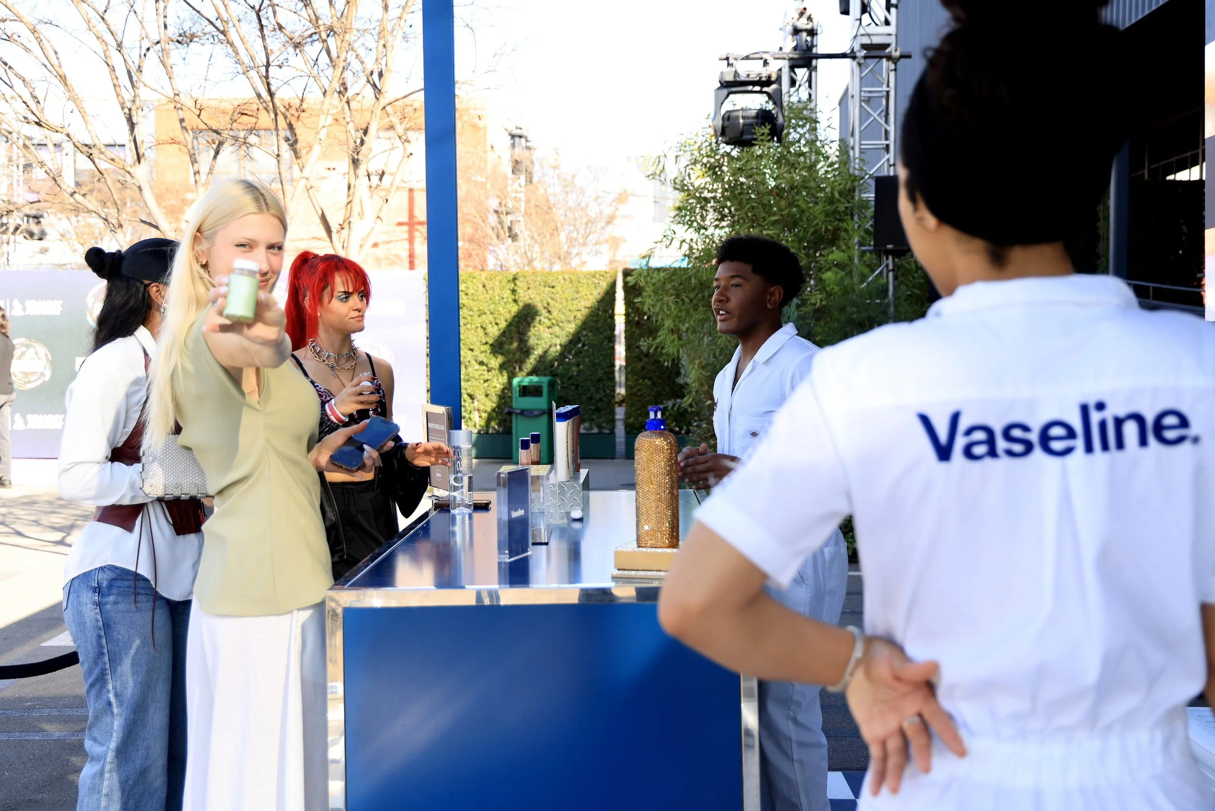 People at a Vaseline booth outdoors, with one person in a white uniform and others in casual clothing, engaging in a conversation, with skincare products on display and a sunny day background.