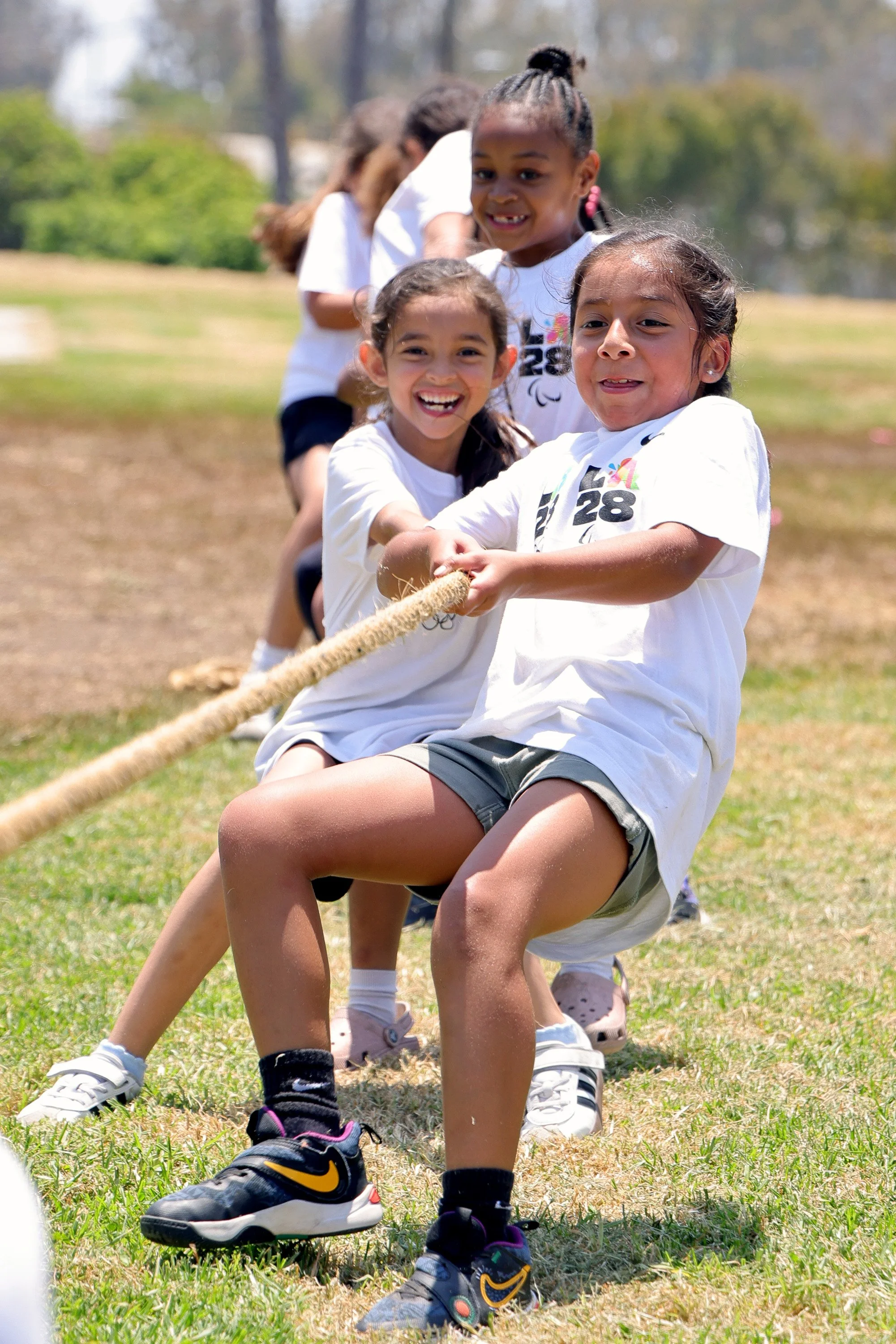 Children participating in a tug-of-war game outdoors on a grassy field on a sunny day, smiling and pulling a rope.
