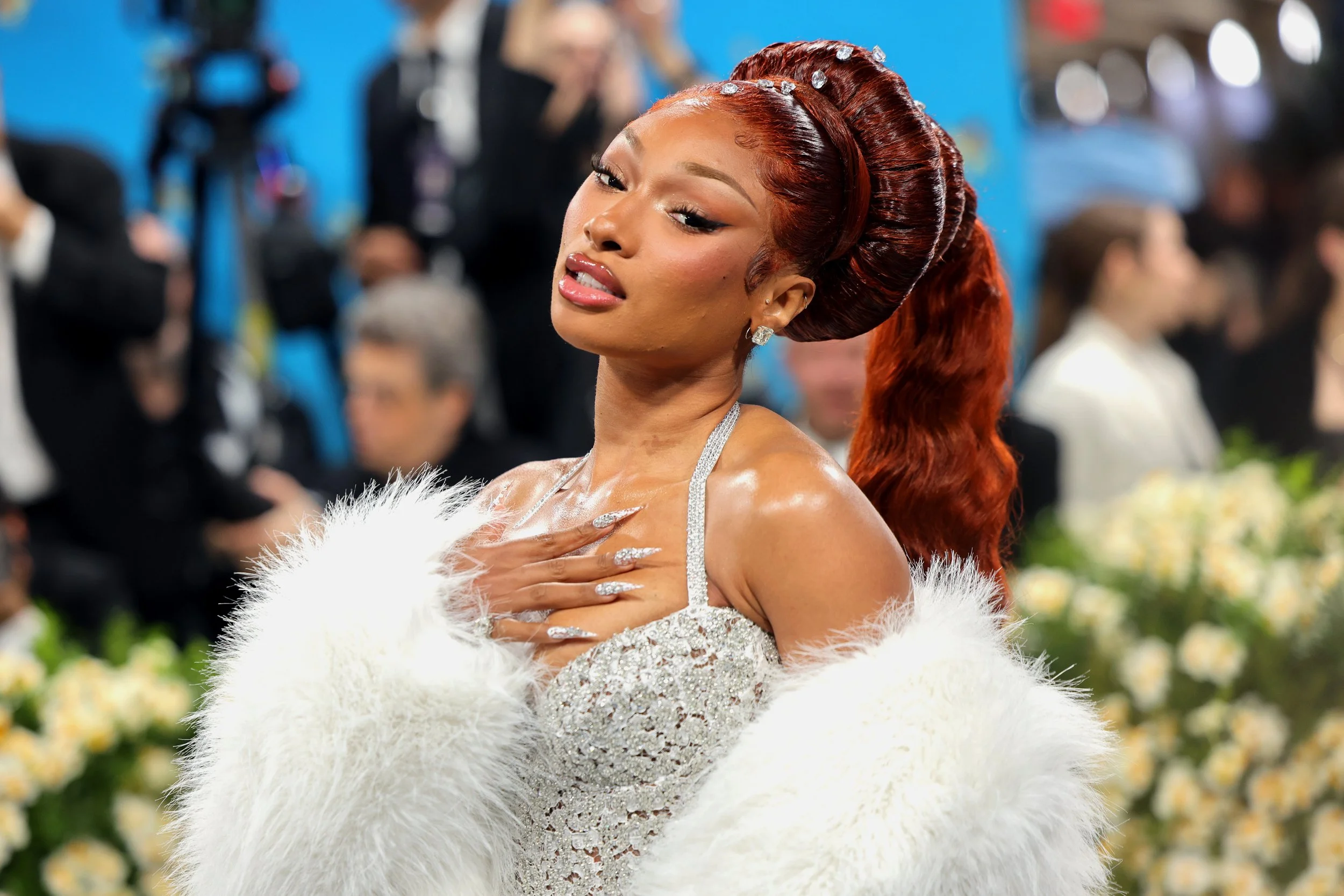 A woman with red hair styled in an elegant updo and long waves, wearing a sparkling gown and a white fur stole, posing at a formal event with a blurred background of other attendees and cameras.