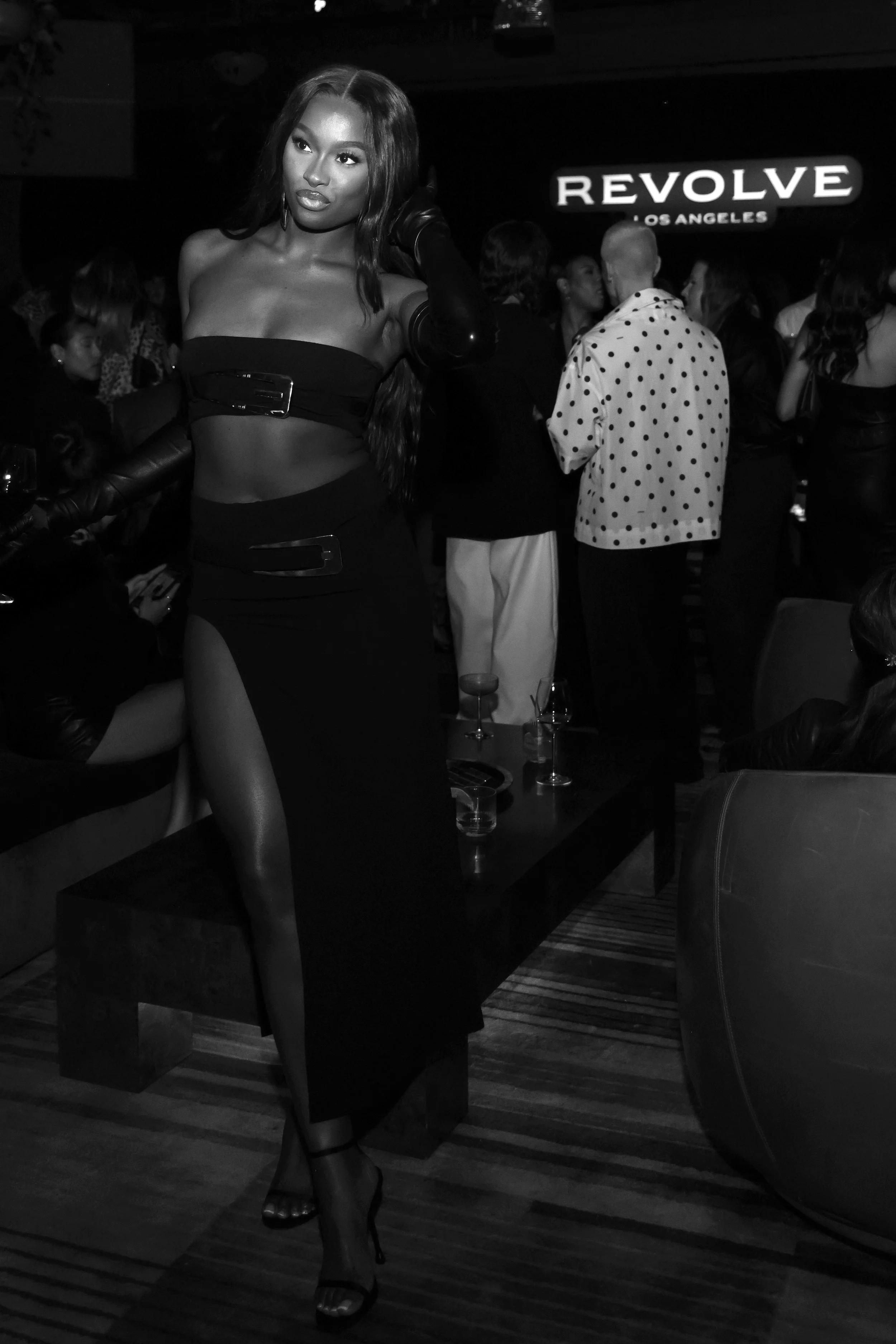 A woman in a revealing black dress posing at a nightclub with a sign that says 'REVOLVE LOS ANGELES' in the background.