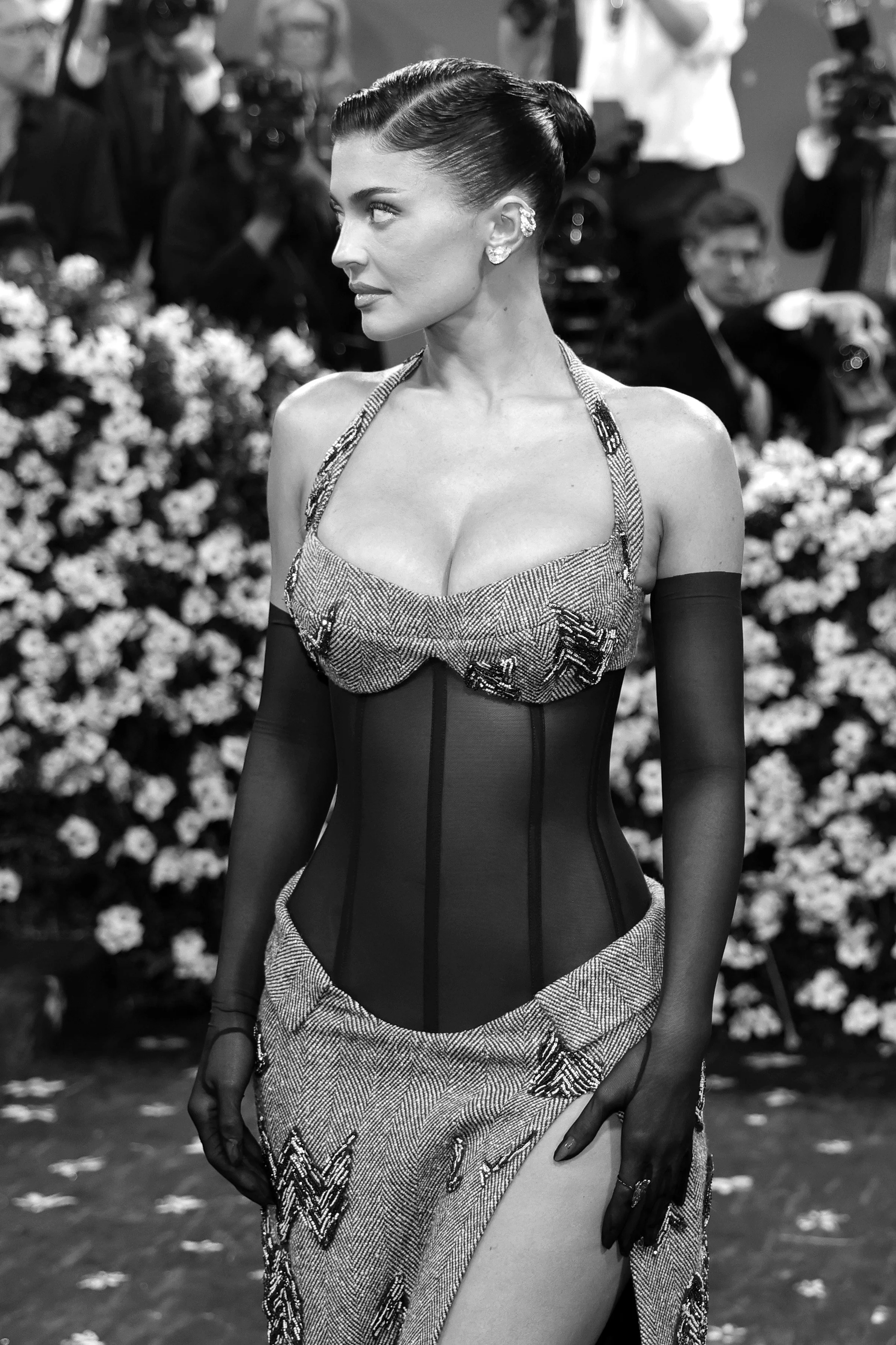 A woman in an elegant dress with a fitted bodice, gloves, and vintage hairstyle, surrounded by photographers and flowers at a formal event.