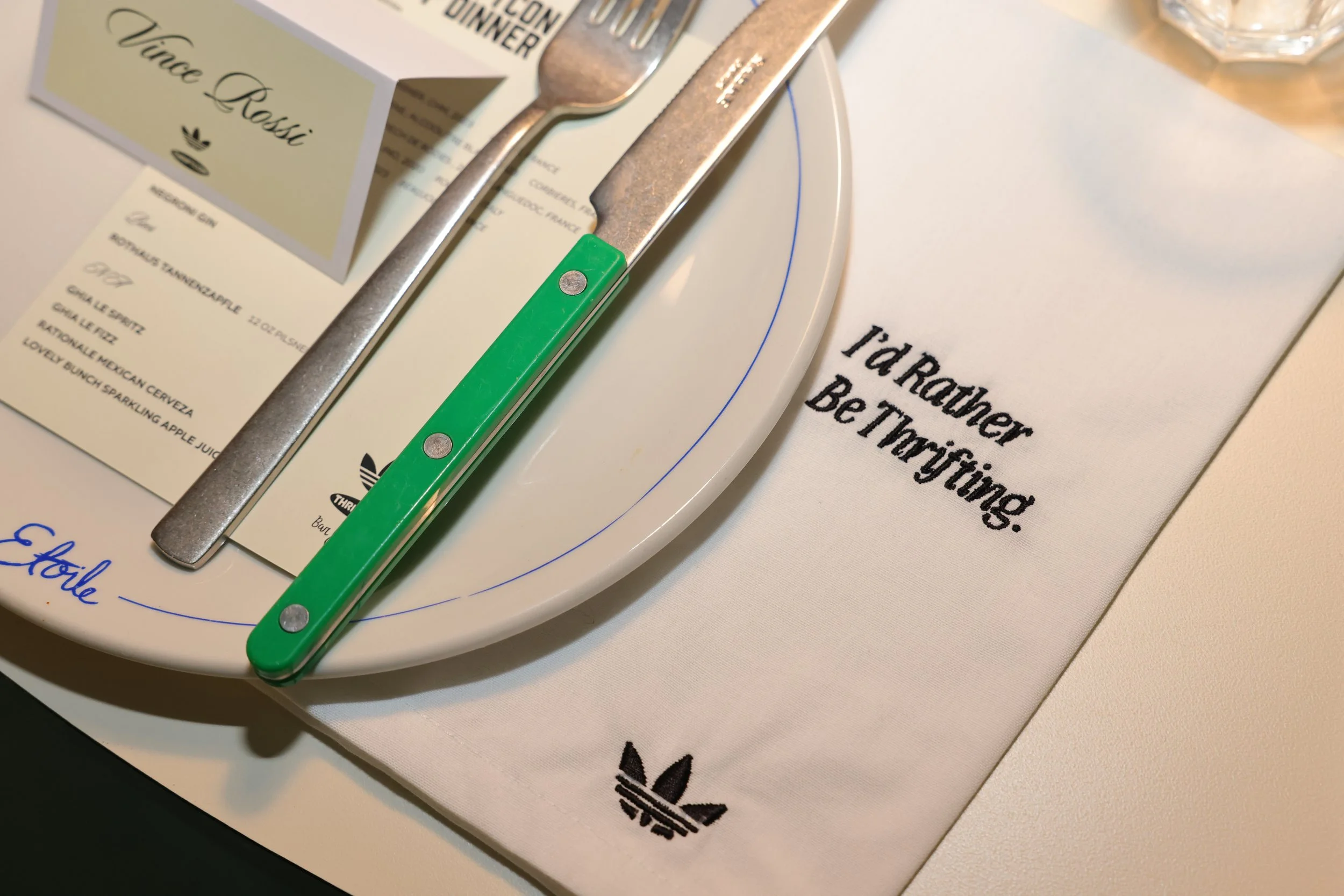 A dining table setting with a white plate, metal knife and fork, a printed menu, a napkin with the message 'I'd rather be Tying.' embroidered on it, and a card with the name 'Vince Rossi.' The items are arranged on a white surface.