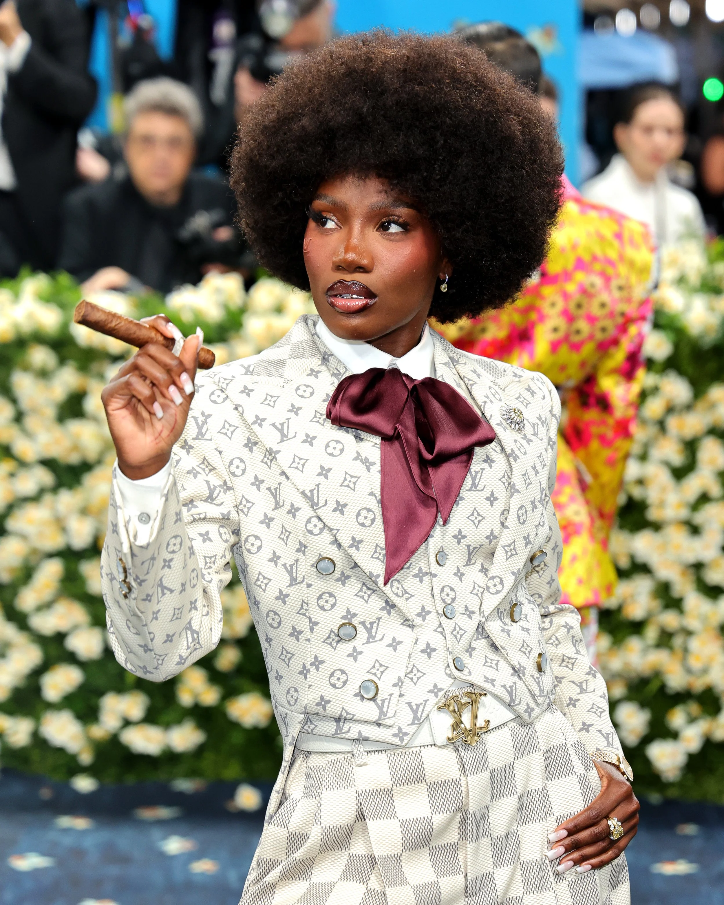 A woman with a large afro hairstyle, wearing a designer Louis Vuitton patterned suit with a red bow tie, holding a cigar, and standing in front of a background of flowers.