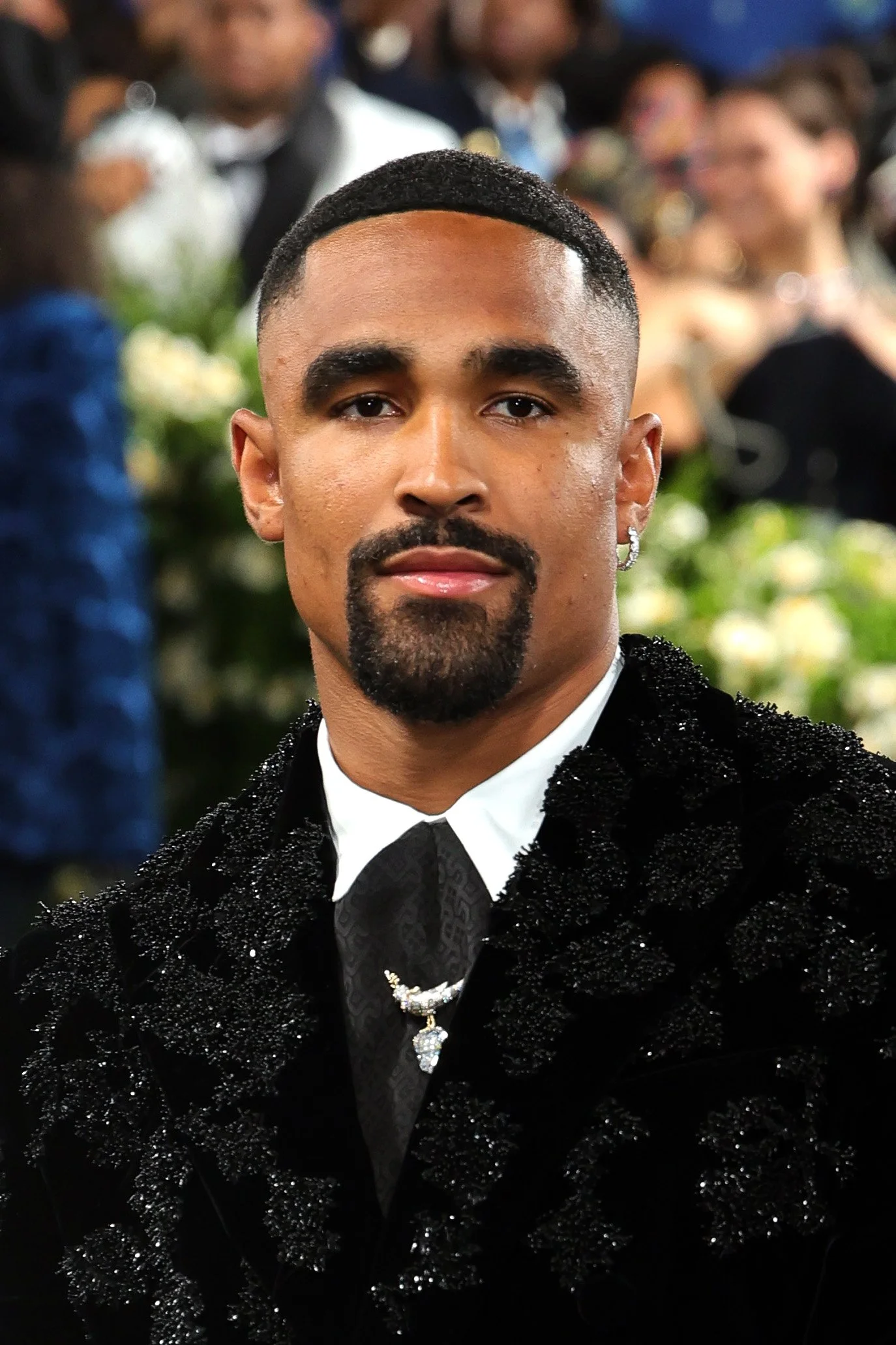 A man with a goatee and mustache, short styled hair, wearing a black, beaded, velvet jacket, white shirt, black tie, and jewelry, sitting at a formal event with blurred people and floral arrangements in the background.