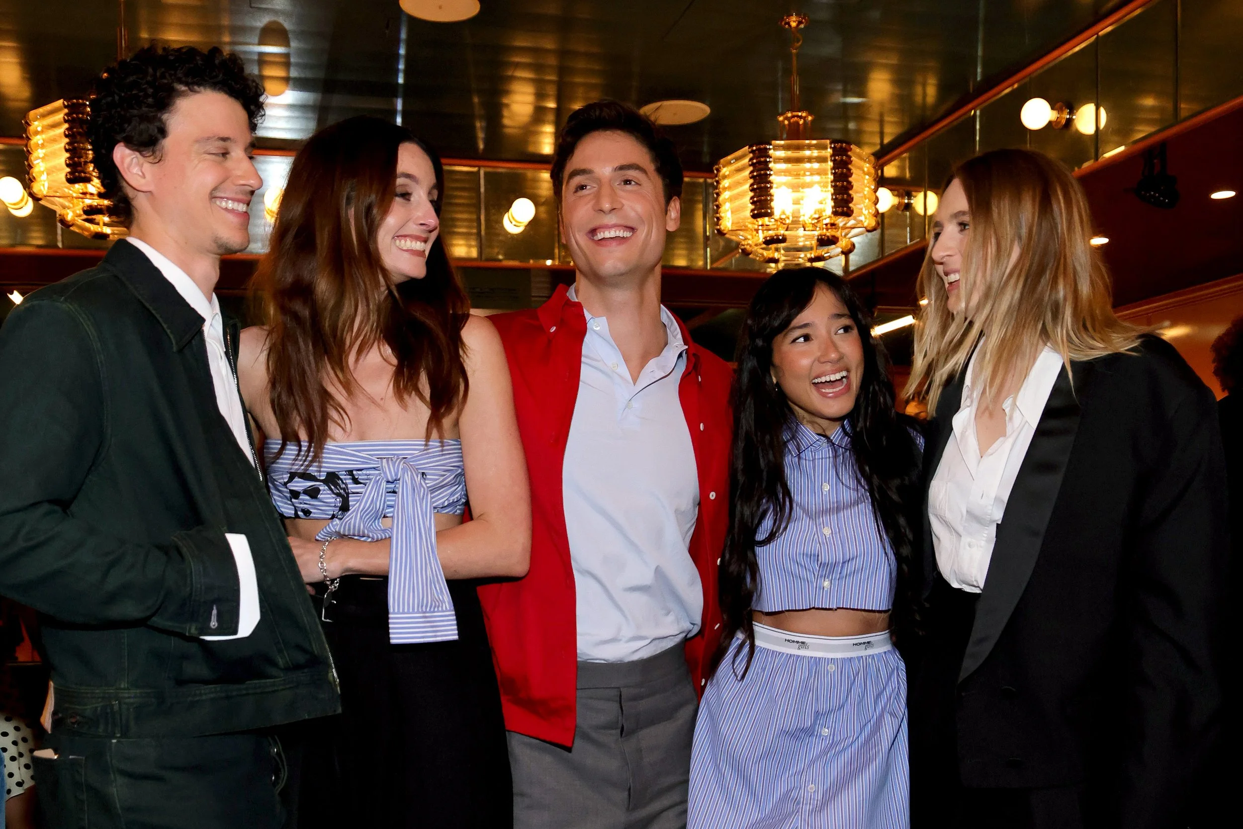 Group of five young adults smiling and chatting in a warmly lit indoor setting, likely a restaurant or bar.