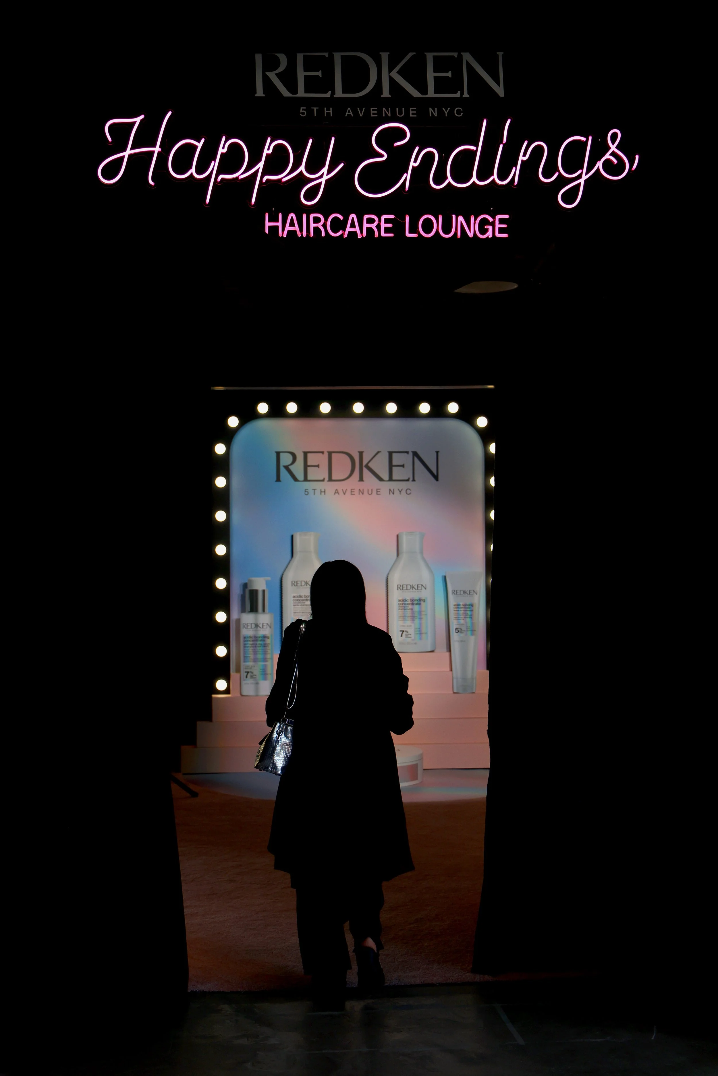 Silhouette of a woman walking through a dark doorway into a brightly lit redken haircare lounge display with various product bottles and a pink neon sign that reads 'Happy Endings' with smaller text 'HAIRCARE LOUNGE' underneath.
