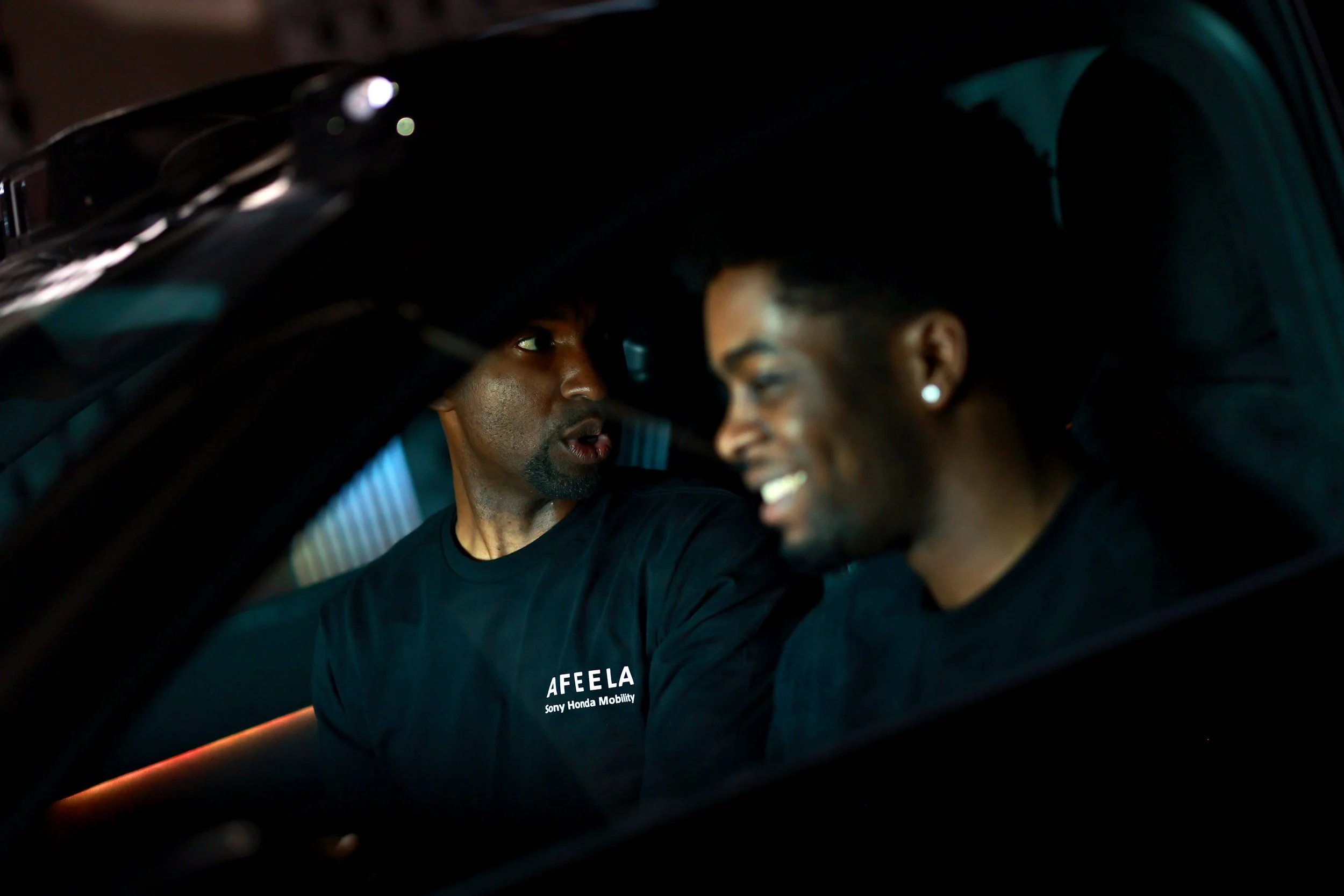 Two men sitting inside a dark car chatting and smiling at each other, one of them wearing a black T-shirt with white text.