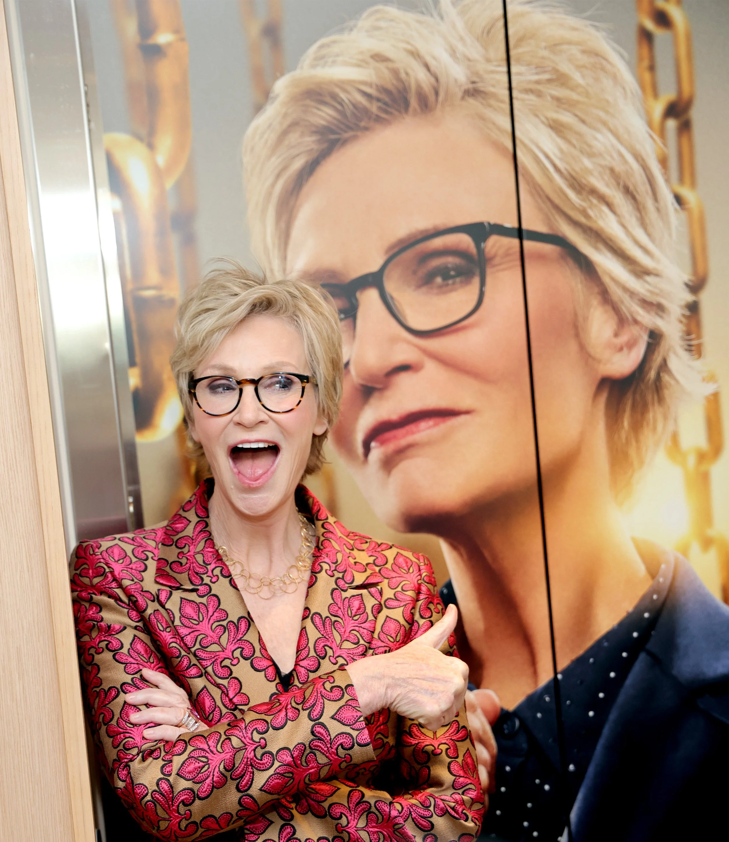 A woman with short blonde hair, wearing glasses, a pink and brown floral blazer, and gold jewelry, standing in front of a large poster of herself, smiling with her mouth open, crossing her arms and pointing at her poster.
