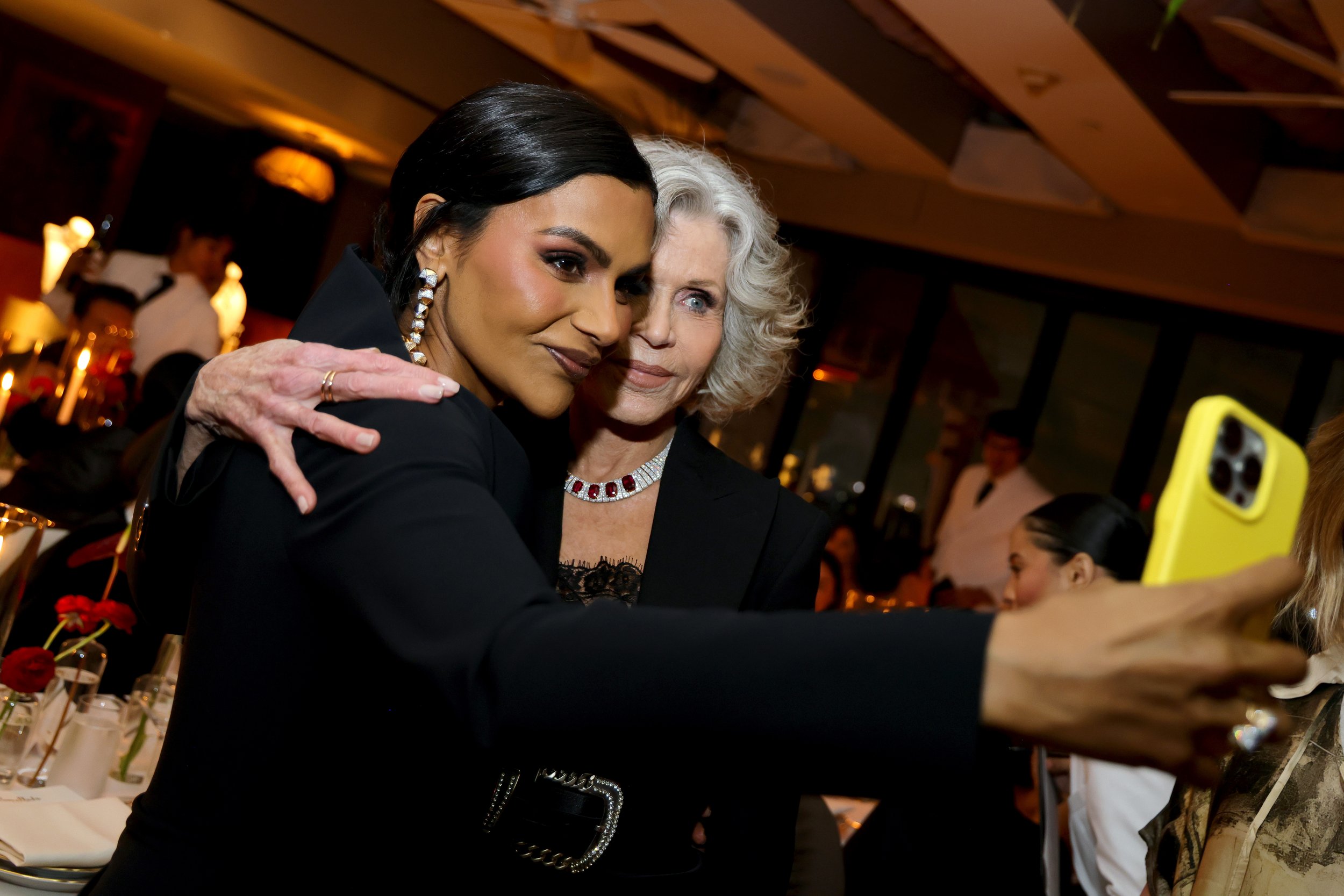Two women taking a selfie together at a formal event, one woman with dark hair and jewelry, the other with white hair and a necklace, in a warmly lit dining area.