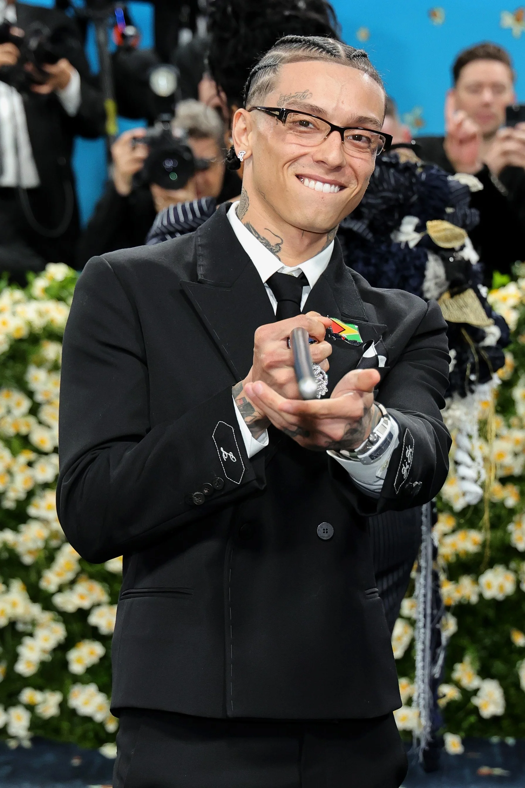 A smiling man with tattoos, glasses, and braided hair, dressed in a black suit, posing at a formal event with photographers and flowers in the background.