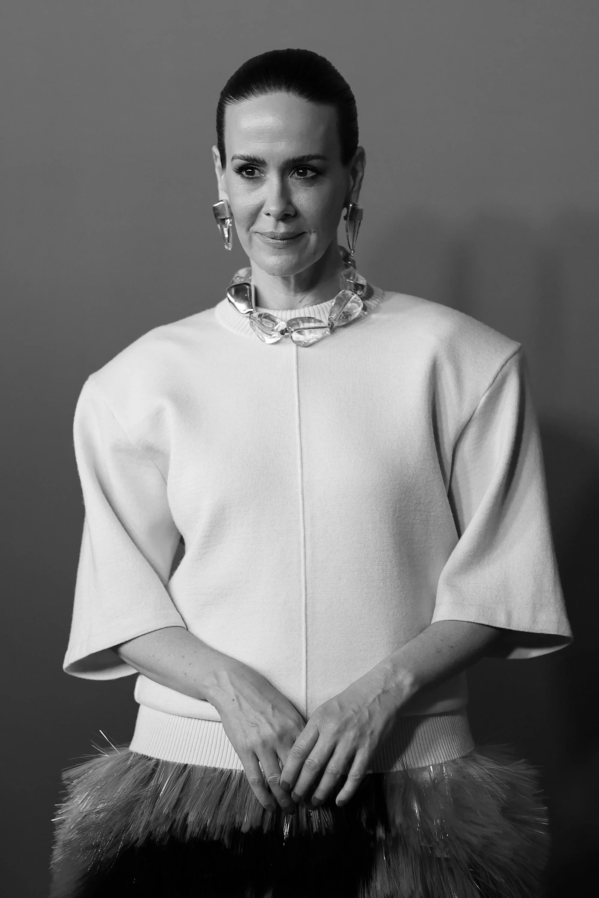 Black and white photo of a woman with dark hair slicked back, wearing a loose-fitting top, large jewelry, and holding her hands together in front of her.
