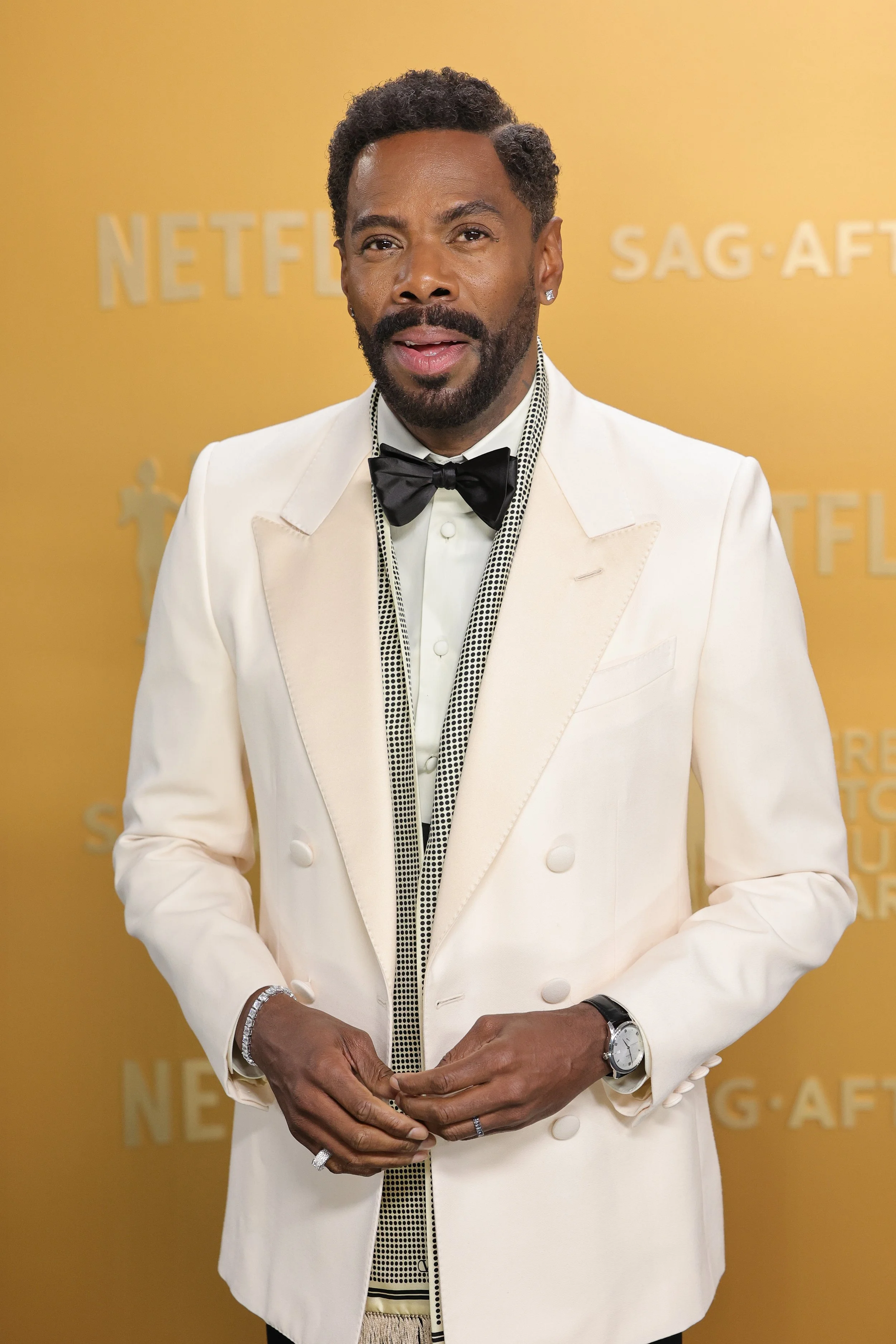 Man wearing a white tuxedo with a black bow tie, standing in front of a yellow background with 'Netflix', 'SAG-AFTRA' and 'TUD' text.