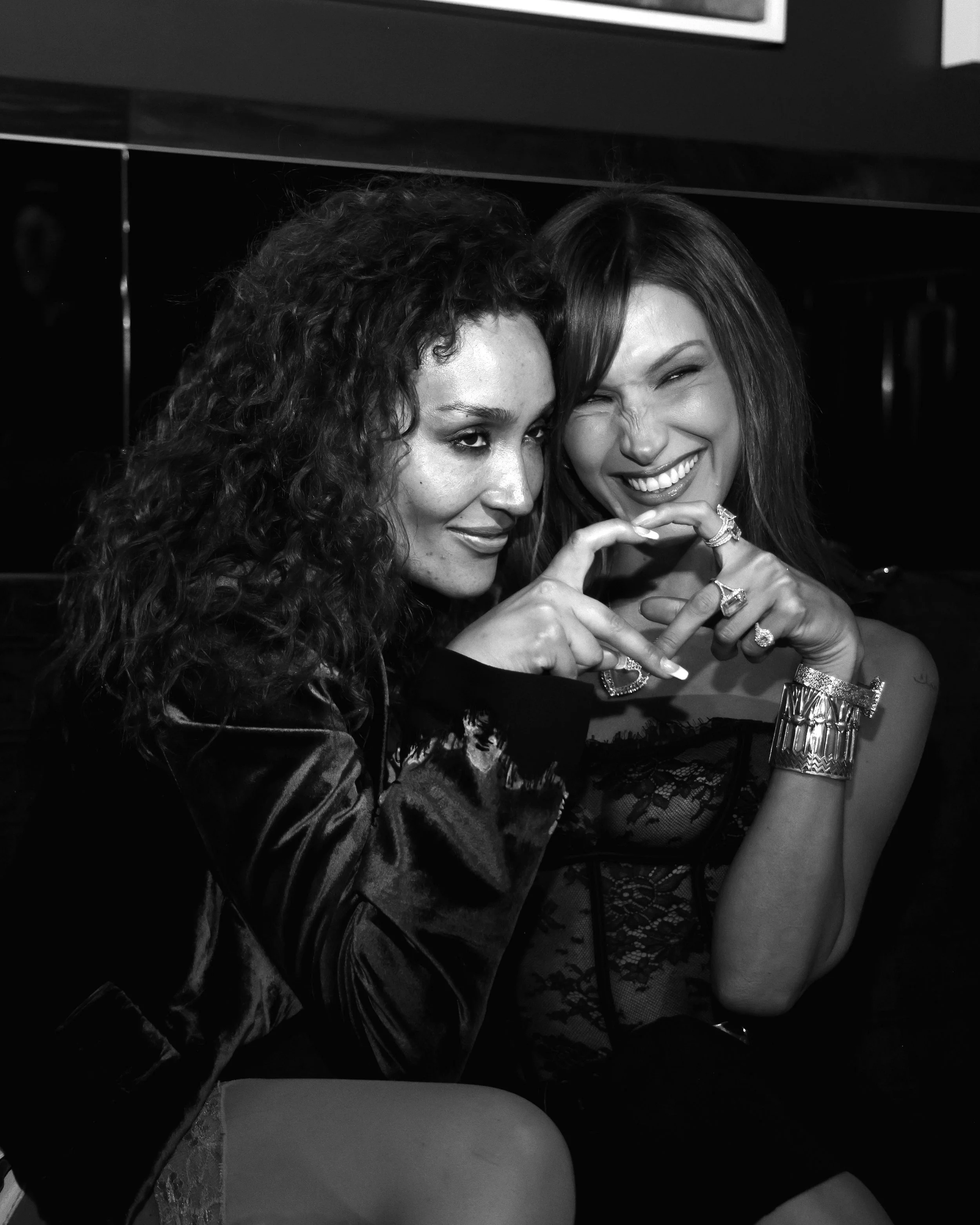Two women with curly and straight hair smiling and making a hand gesture together, wearing jewelry and dark clothing.