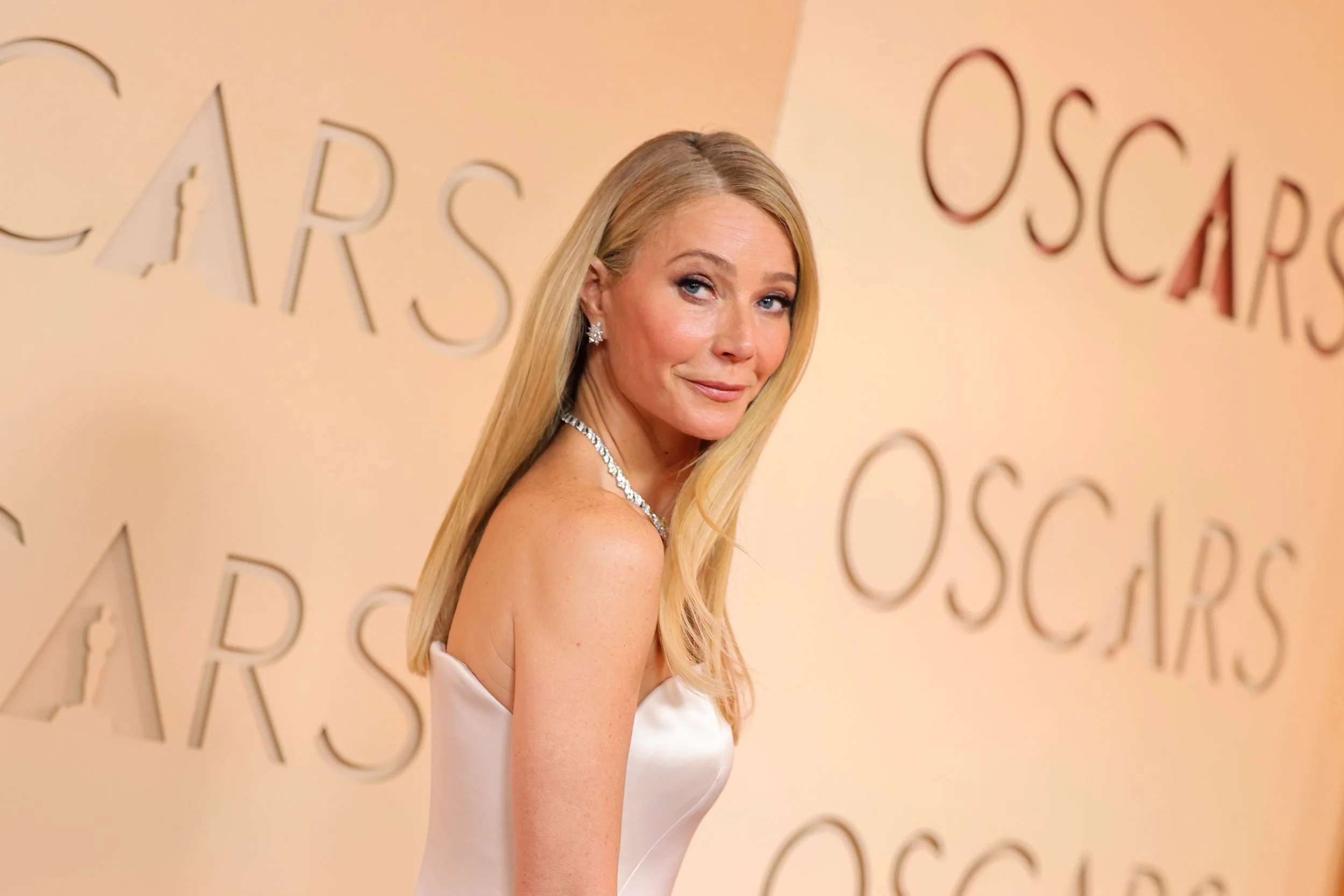 A woman with long blonde hair poses on the red carpet at the Oscars, wearing a strapless white gown and diamond jewelry, with the Oscars logo visible in the background.