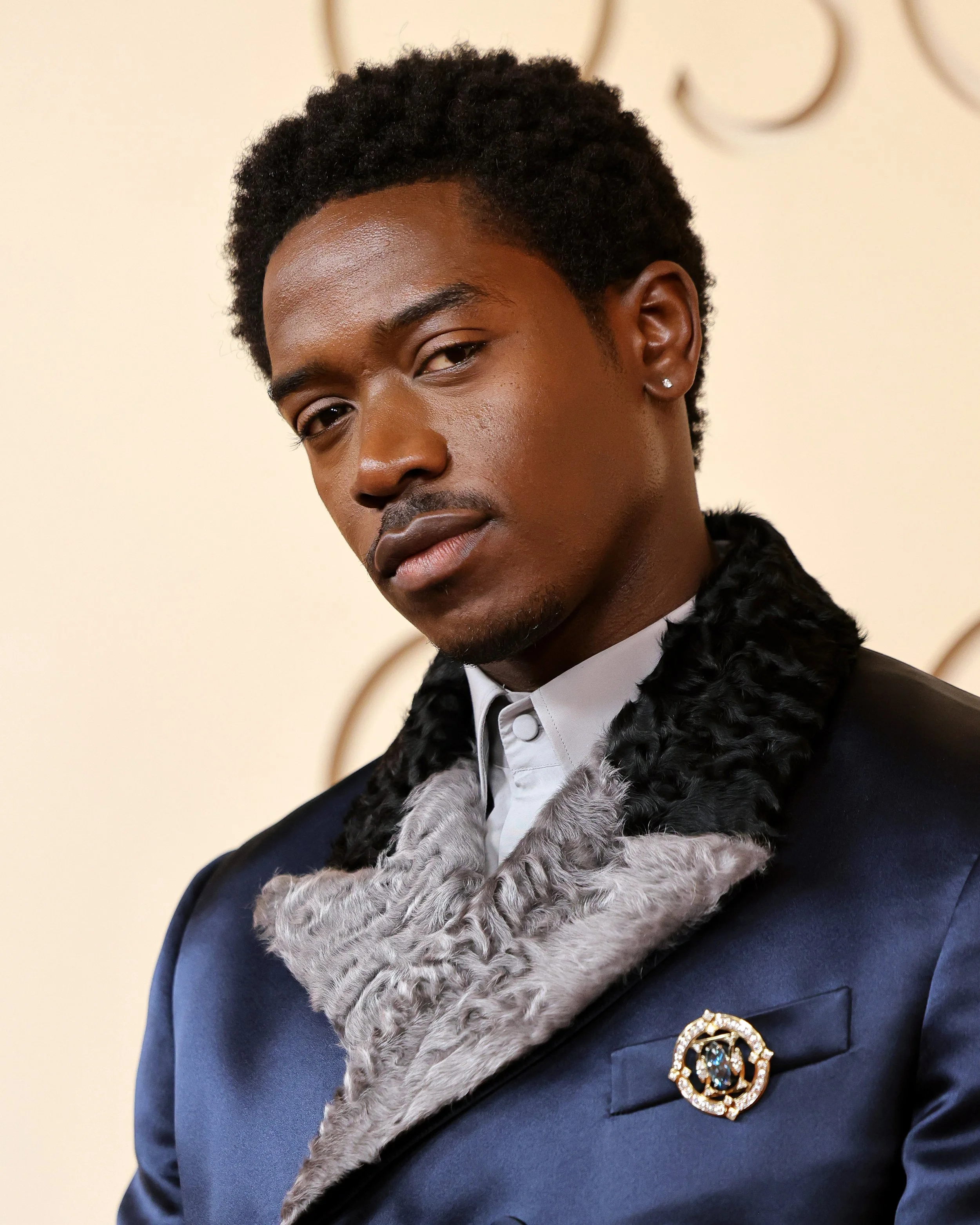 A young man with dark skin and short curly hair wearing a dark blue suit with a fur-lined collar, a white shirt, a brooch, and a small earring on his left ear.