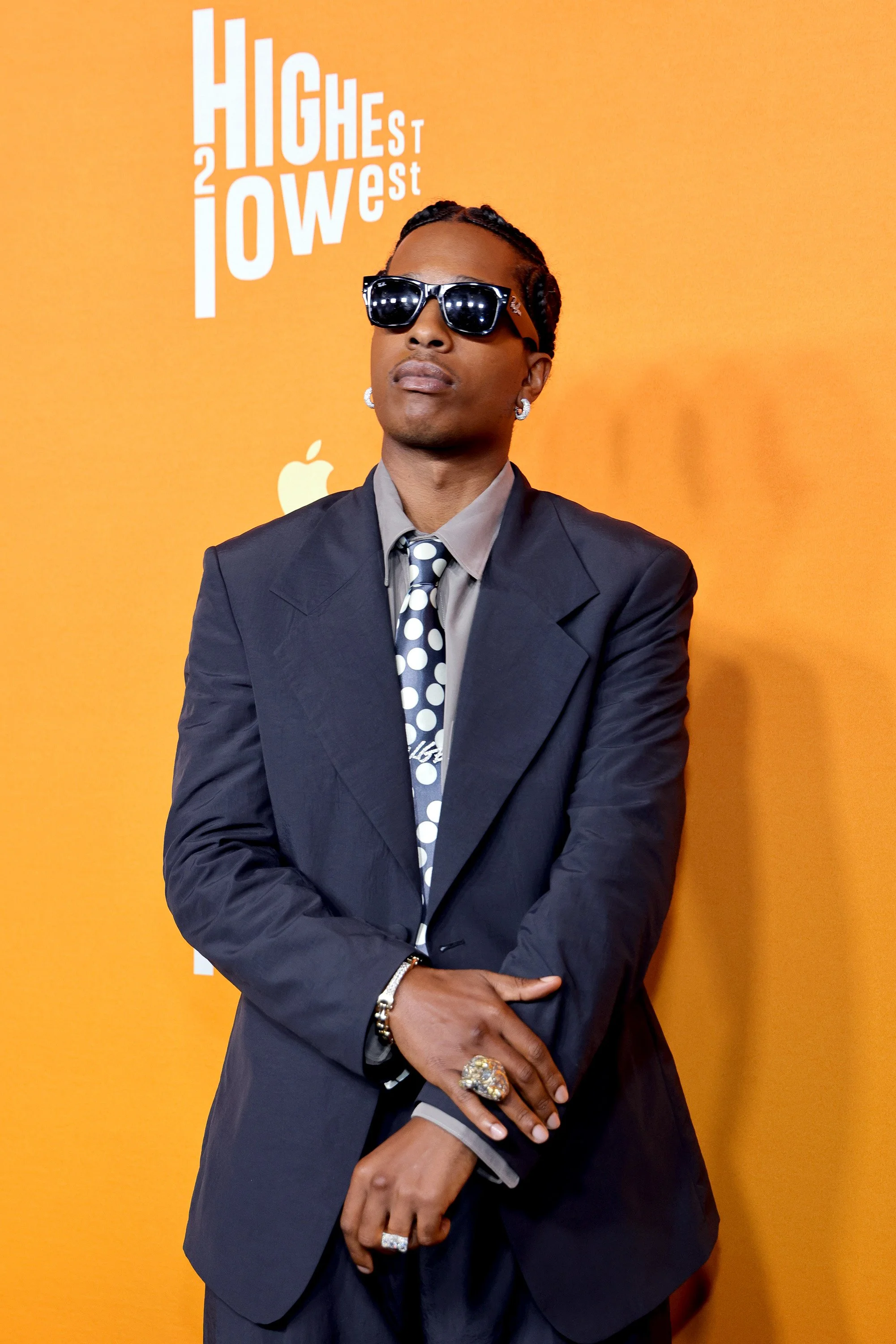 A man wearing sunglasses, a dark suit, and a polka dot tie stands in front of an orange background with white text and a logo. He has earrings and rings.