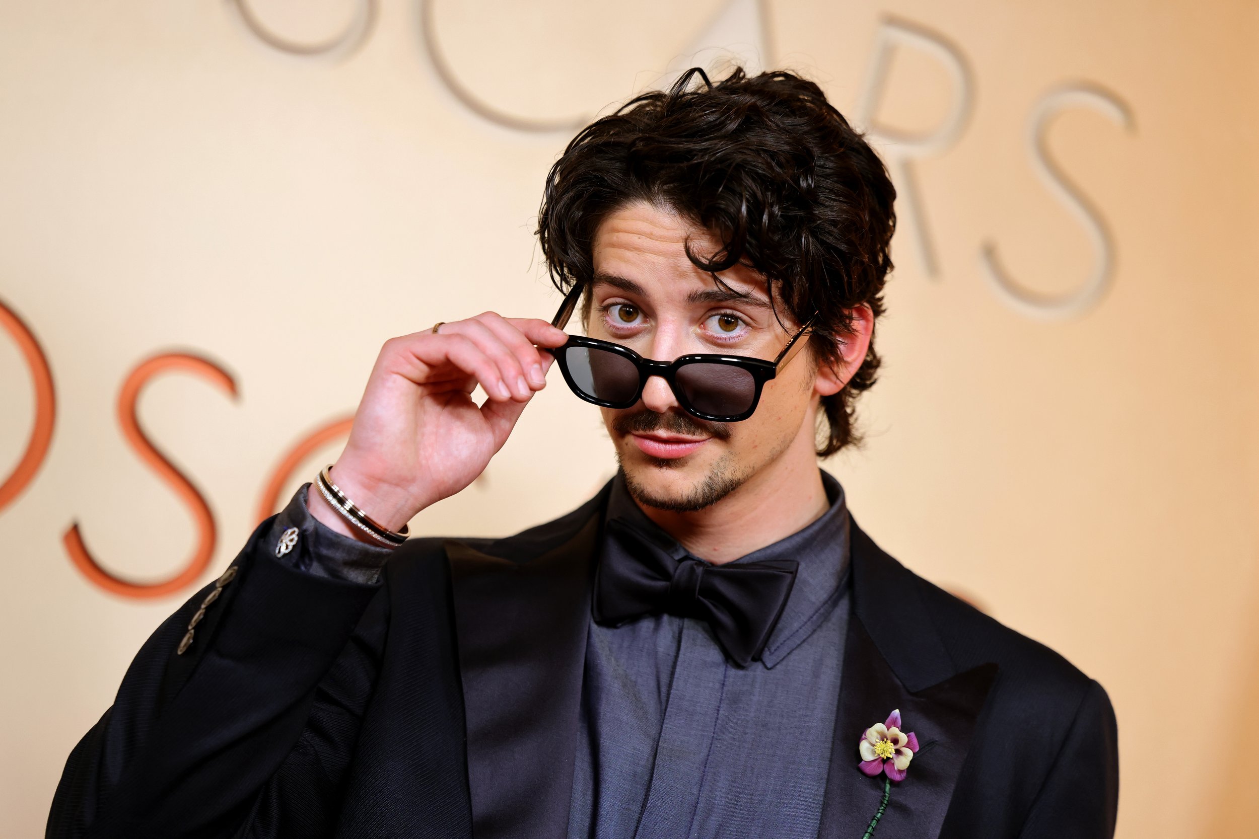 A man with dark curly hair wearing a dark suit and bow tie, with a purple flower boutonniere, holding sunglasses and looking at the camera in front of a beige background with partial text.