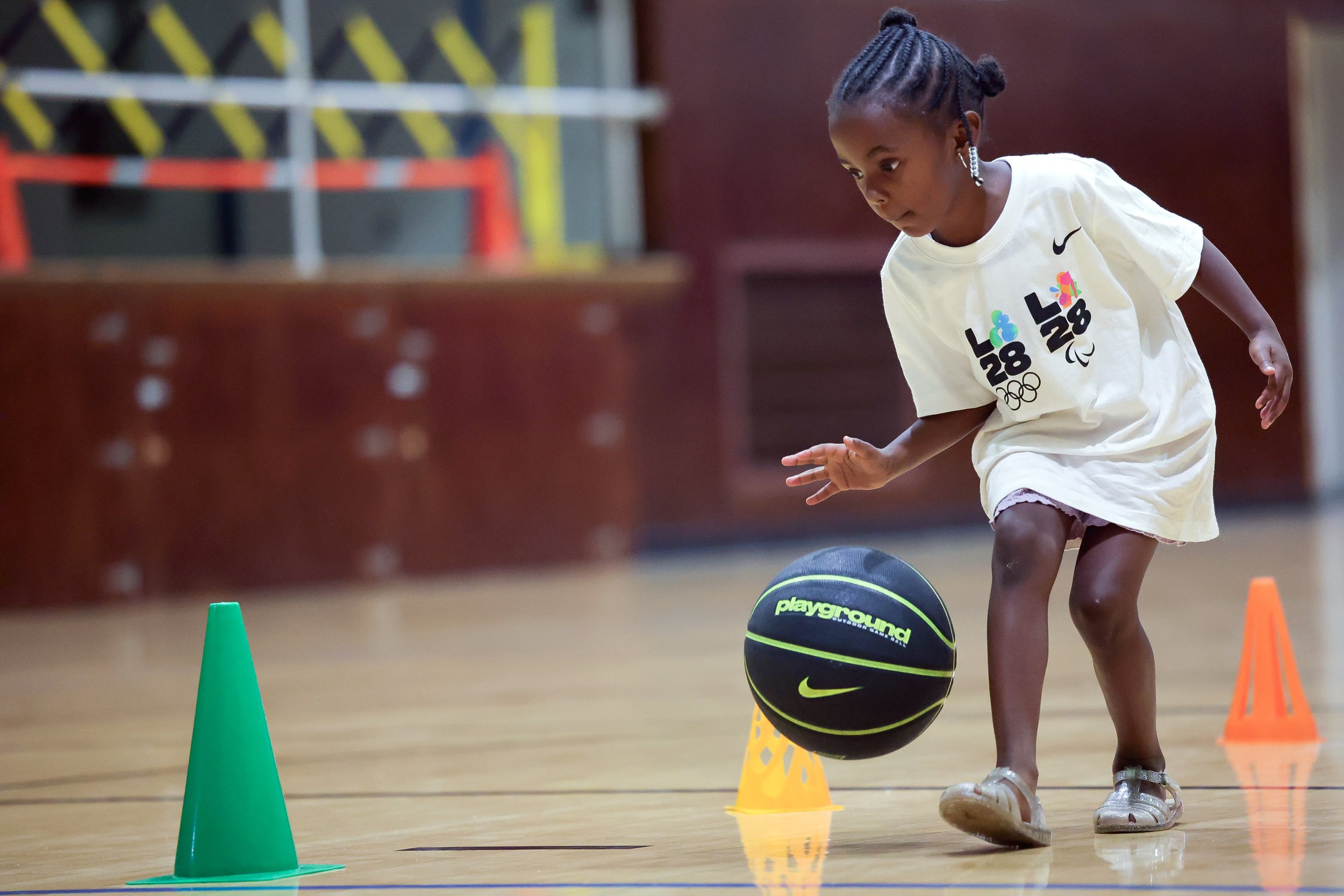 Young girl dribbling a basketball around orange and green cones on a gymnasium floor.