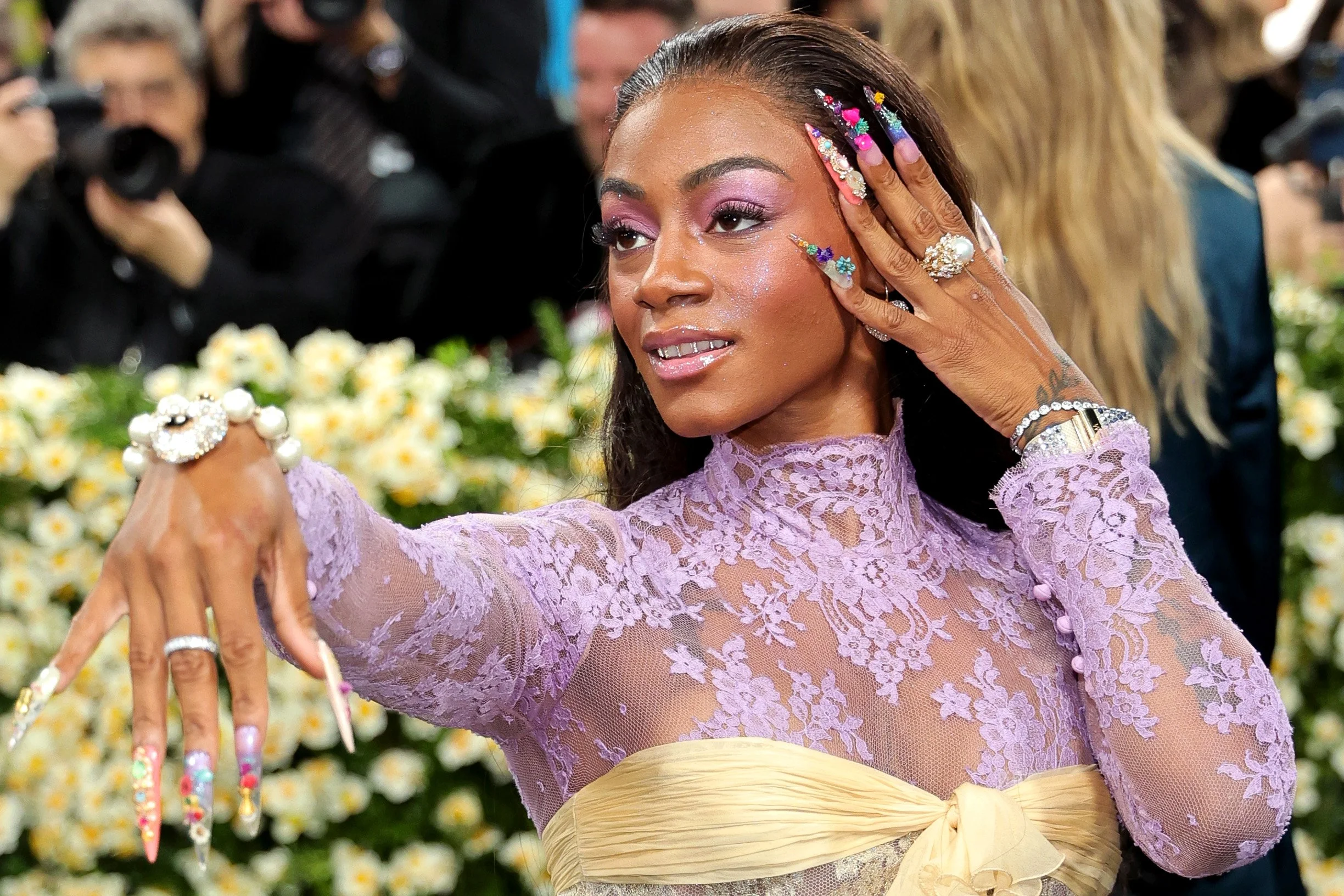 A woman with long dark hair and colorful, decorated nails is posing with her hand near her face. She is wearing a lavender lace top with a yellow ribbon at the waist, and has multiple rings and bracelets. She has pink eyeshadow and glitter on her fac