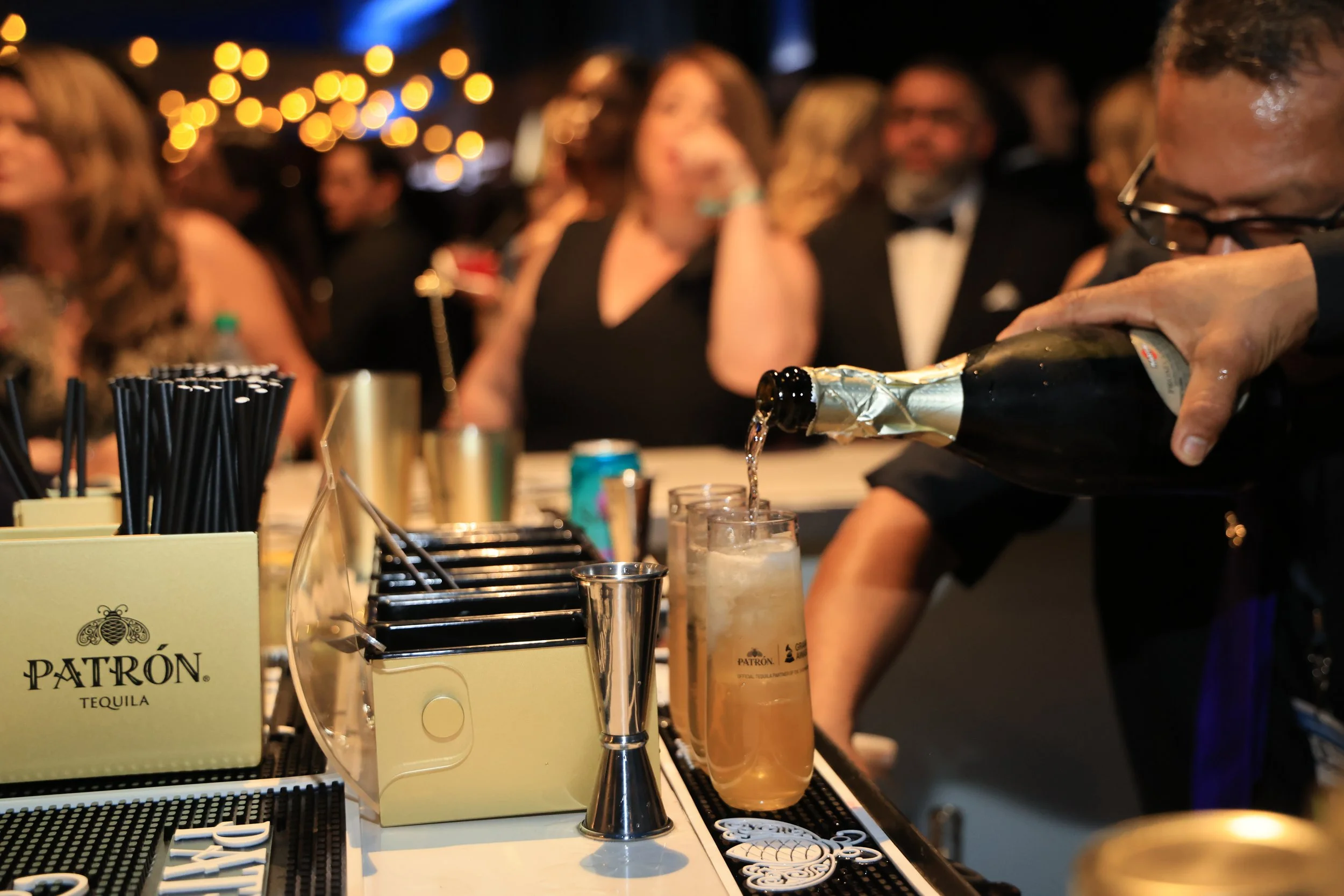 Bartender pouring a drink at a crowded event with guests seated at a bar, illuminated by string lights.