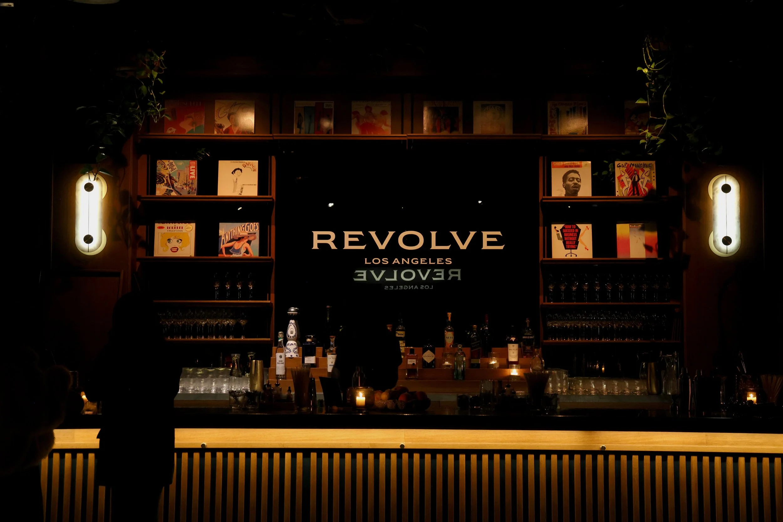 A dimly lit bar with the word 'REVOLVE' and 'LOS ANGELES' on a black background. Shelves with artwork and books flank the bar, with glasses and bottles behind the counter, and a person standing at the bar.
