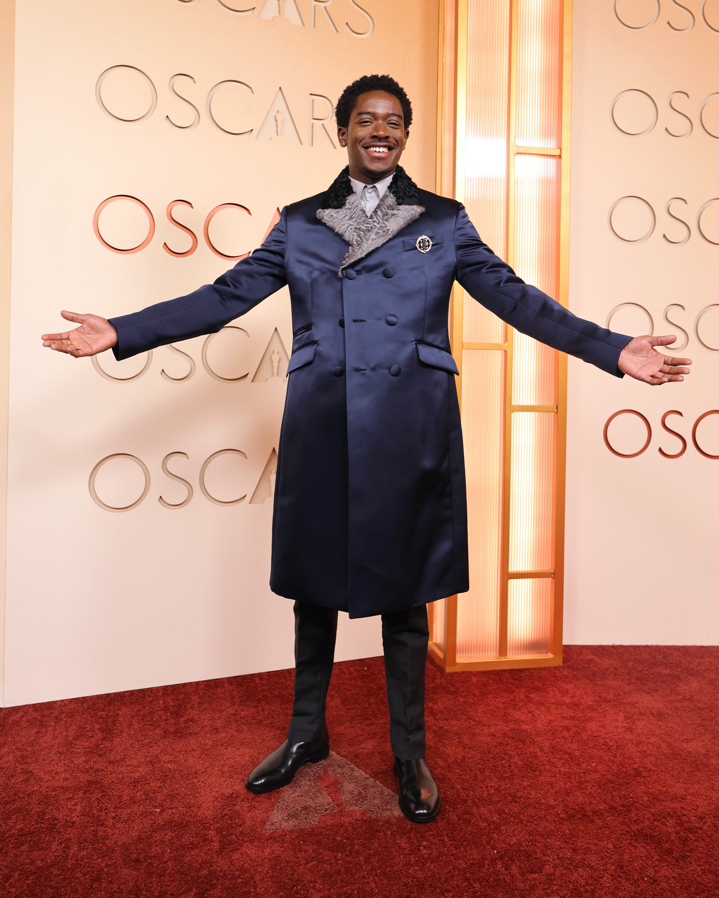 A smiling man standing with arms open in front of a backdrop with the word 'Oscars' repeated in the background at an awards event.