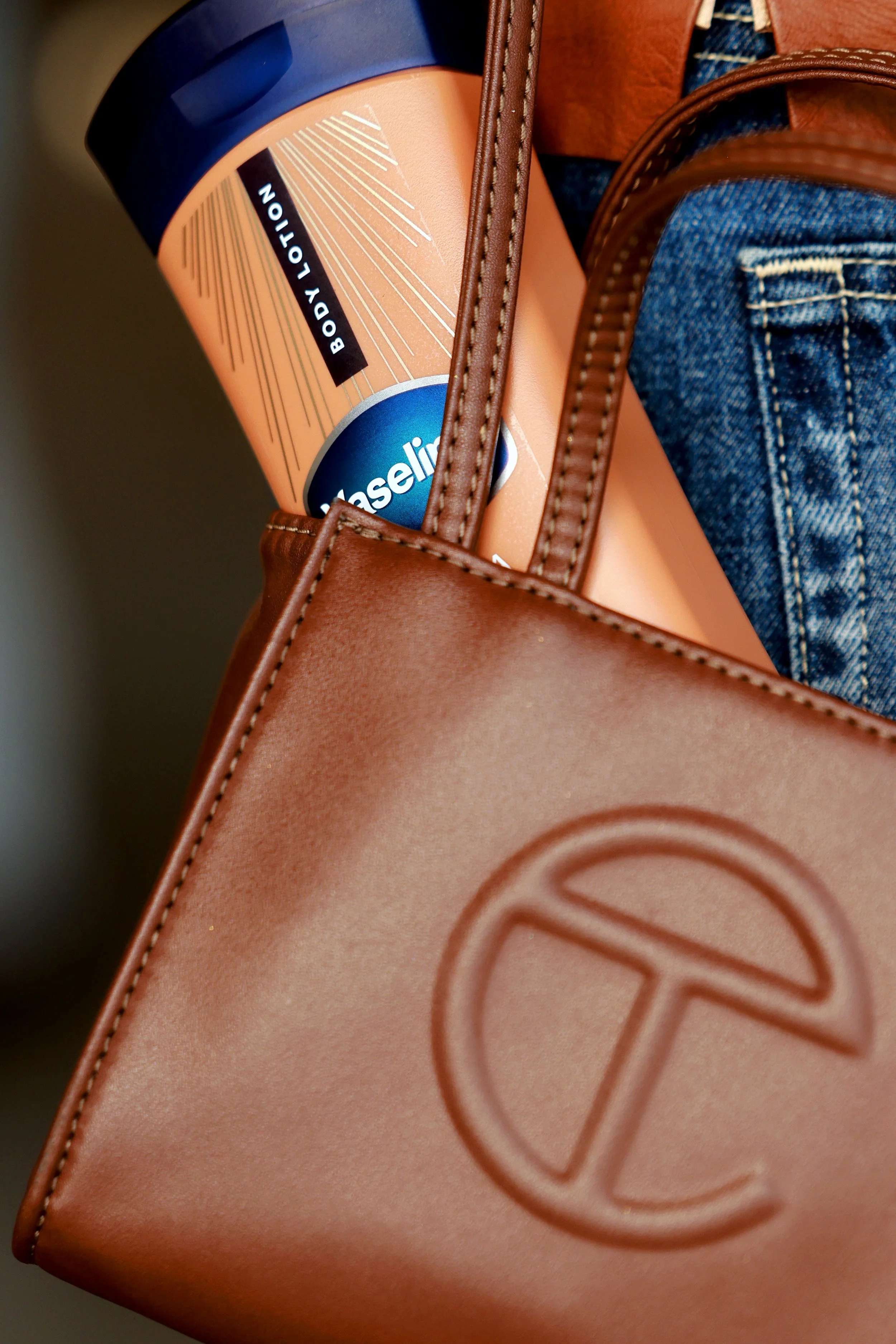 A close-up of a brown tote bag with the logo T on it, containing a tub of Costco Kirkland Body Lotion and some denim jeans.