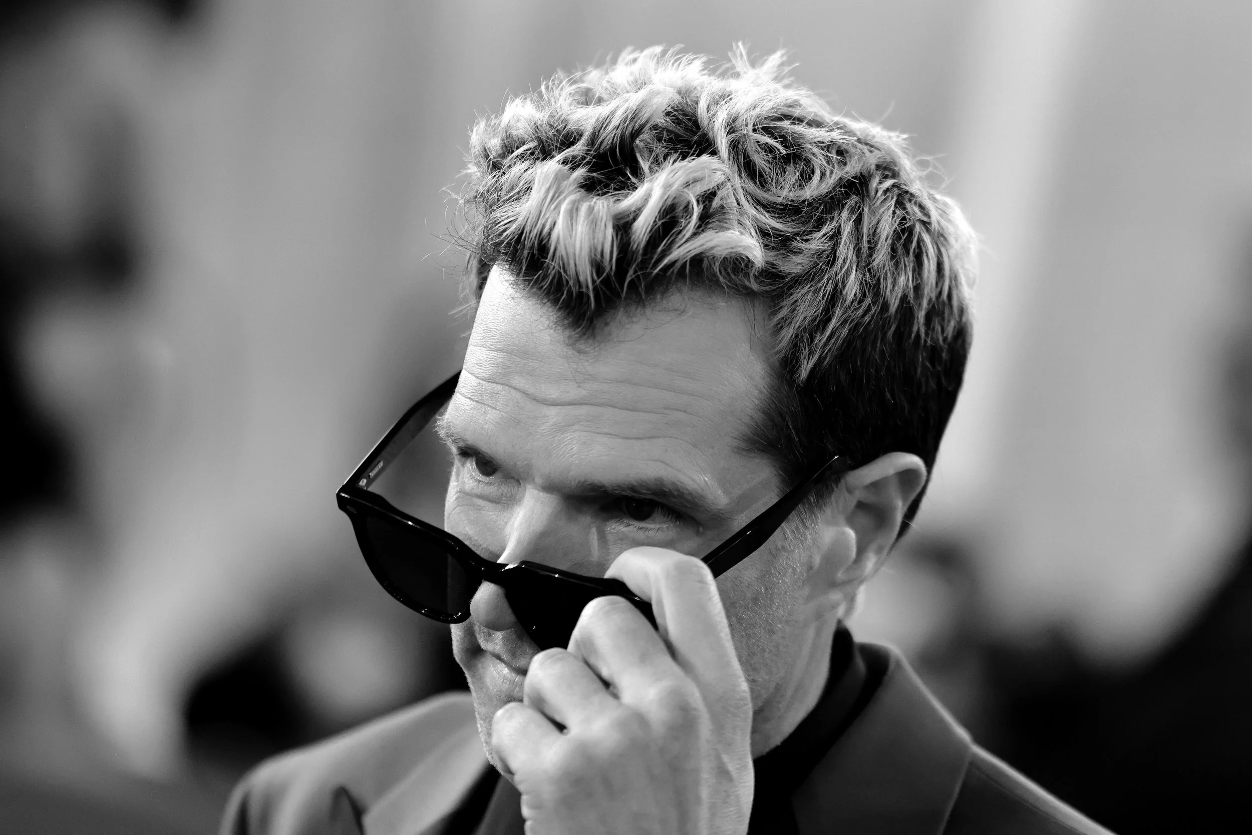 Black and white photo of a man with blonde wavy hair wearing dark sunglasses, looking down and adjusting the sunglasses with his hand.