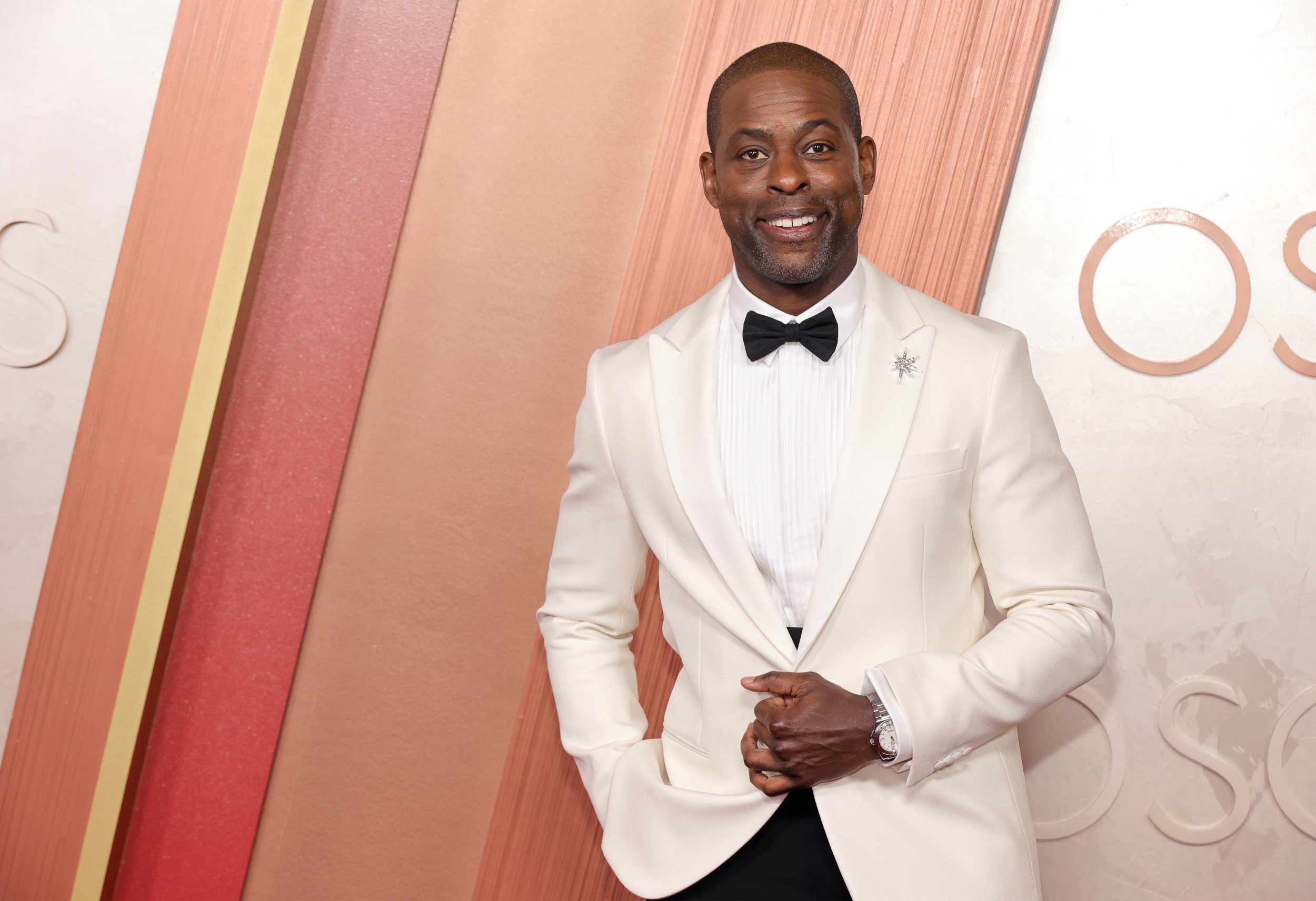 Man in a white tuxedo with a black bow tie standing in front of a colorful background at an event.