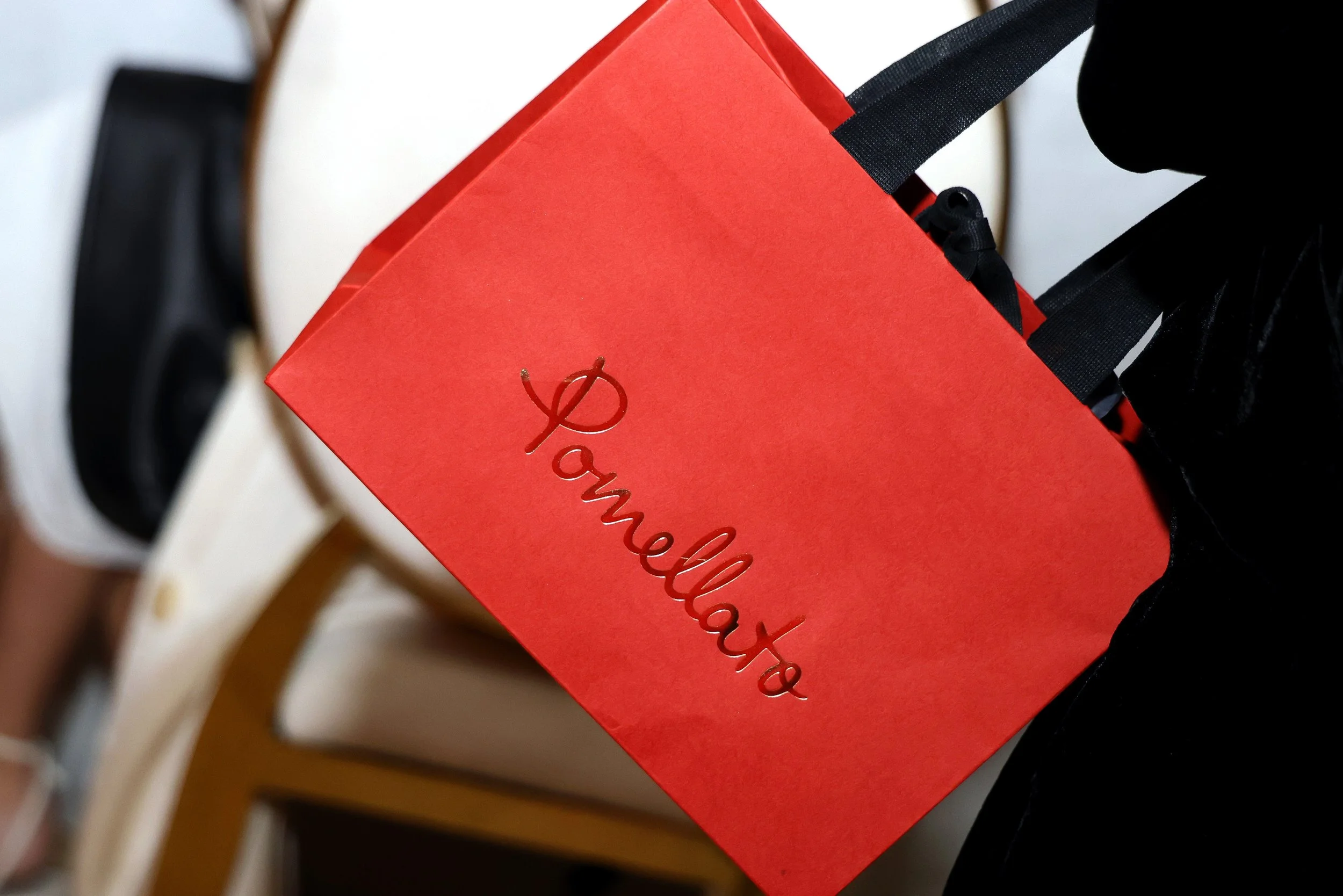 A red gift bag with the word 'Pamellato' embossed on it in cursive, held by someone wearing black clothing, with white chairs in the background.