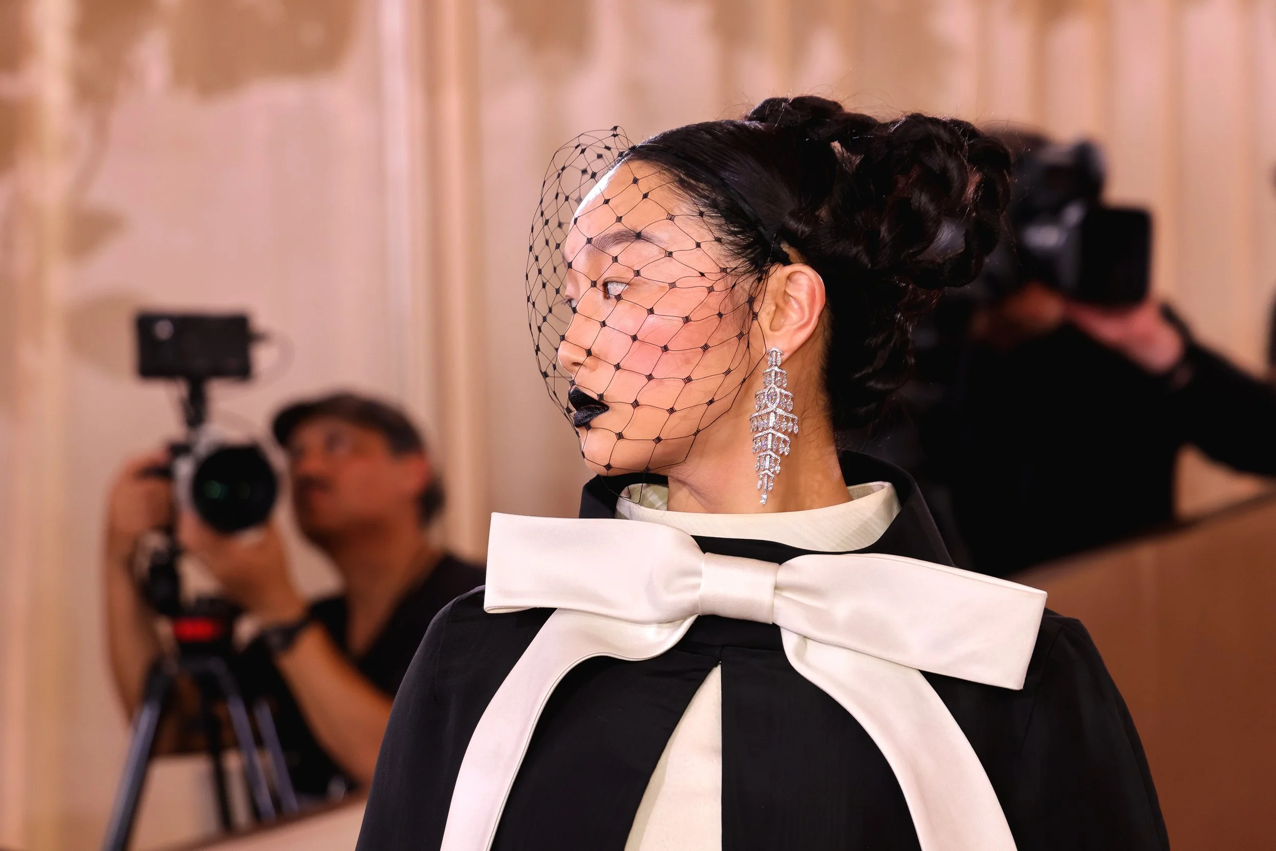 A woman with black hair styled in an elegant updo, wearing a fishnet veil, large chandelier earrings, and black lipstick. She is dressed in a black outfit with a white bow at the collar. Photographers can be seen in the background capturing the momen