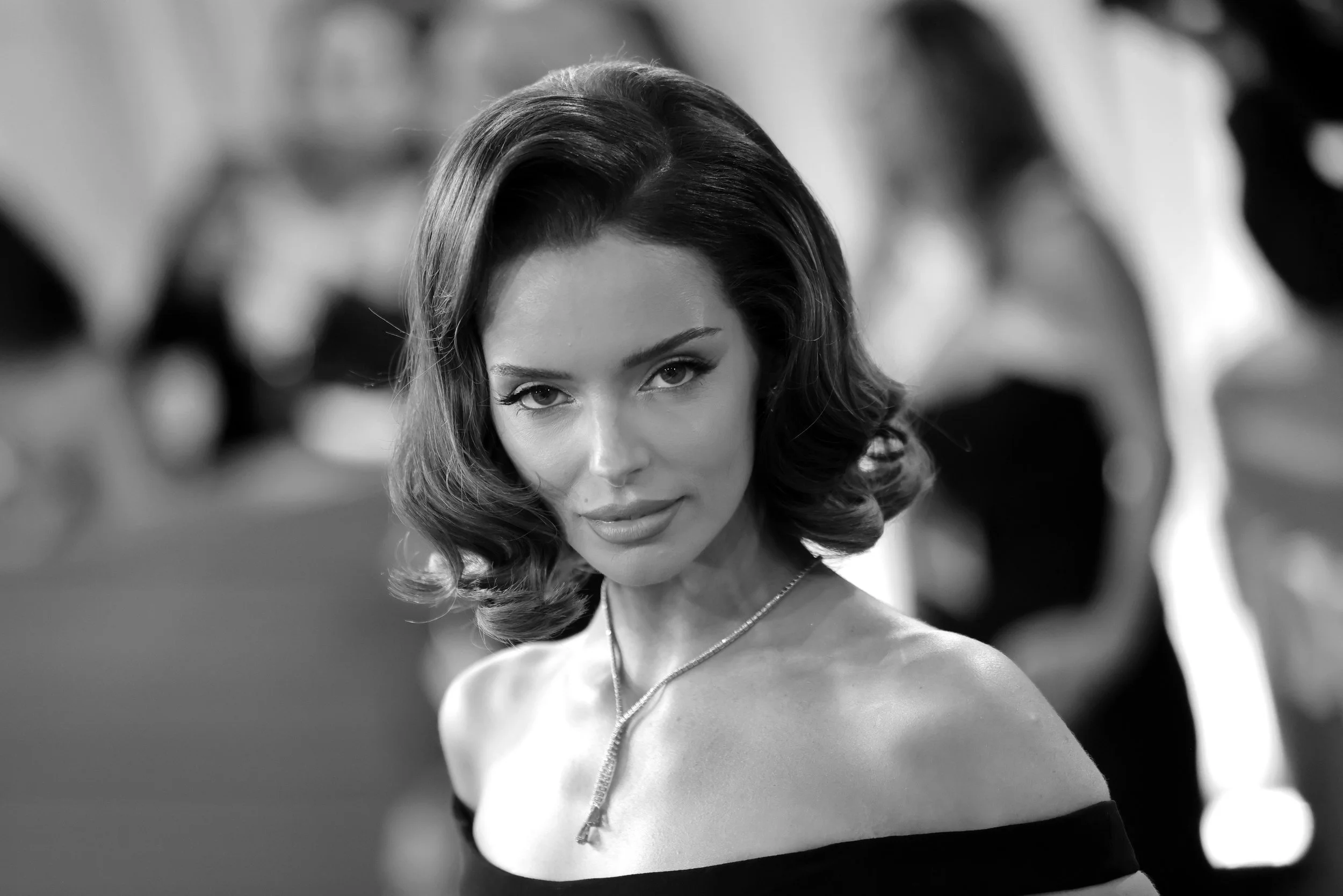 Black and white photograph of a woman with wavy, shoulder-length hair, wearing an off-shoulder top and a necklace, looking directly at the camera with a confident expression.