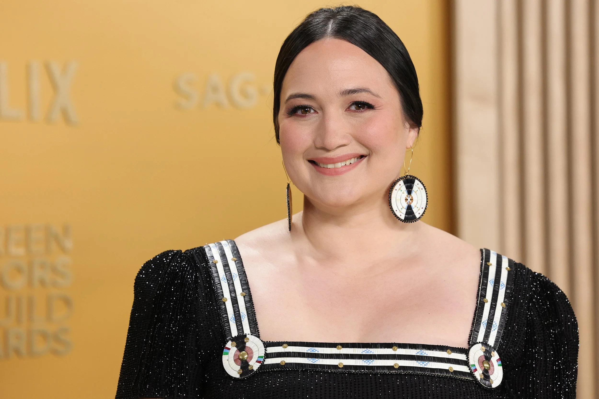 A woman with short black hair, wearing a black dress with silver embellishments, and large circular earrings with a geometric design, smiling at a yellow backdrop with partially visible text.