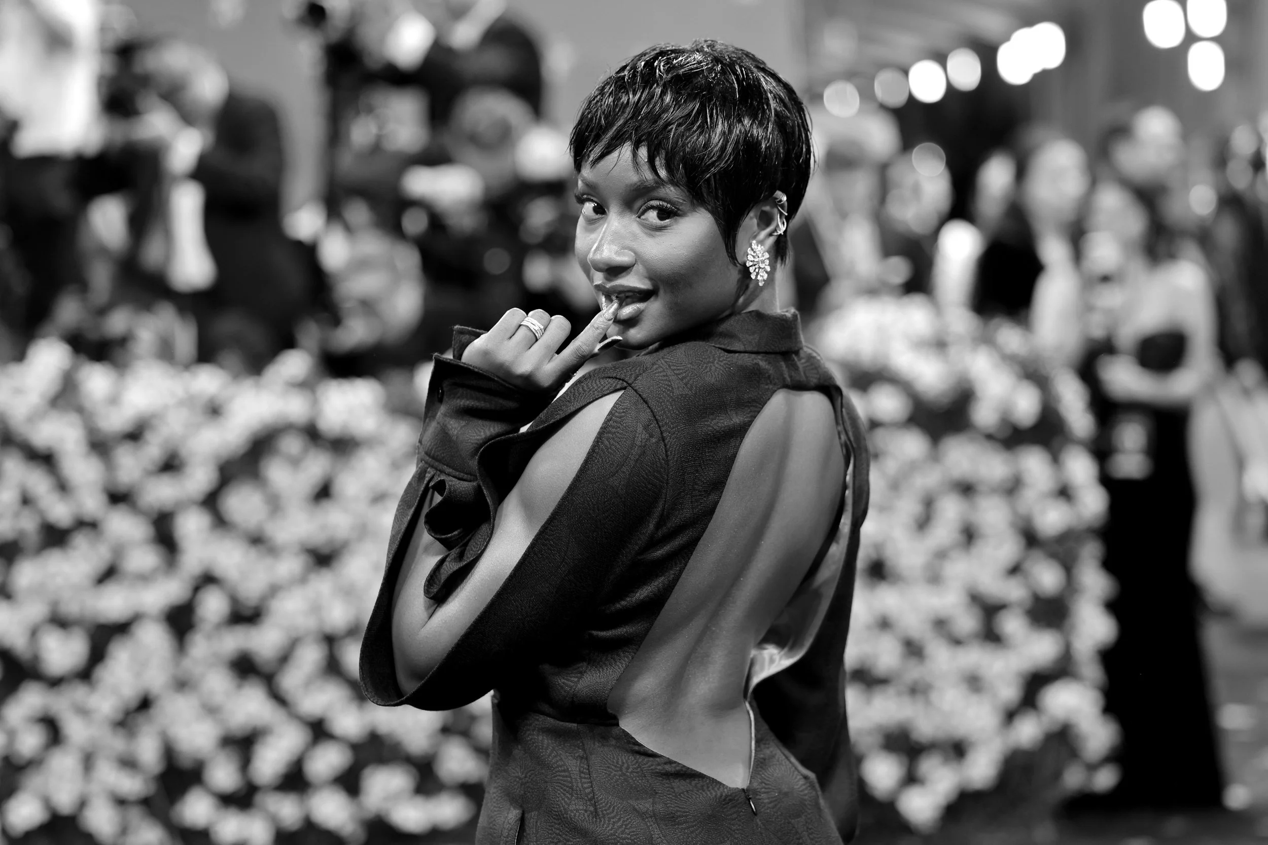 A woman with short hair wearing earrings and a stylish jacket, posing and smiling at a nighttime outdoor event with blurred figures and lights in the background.