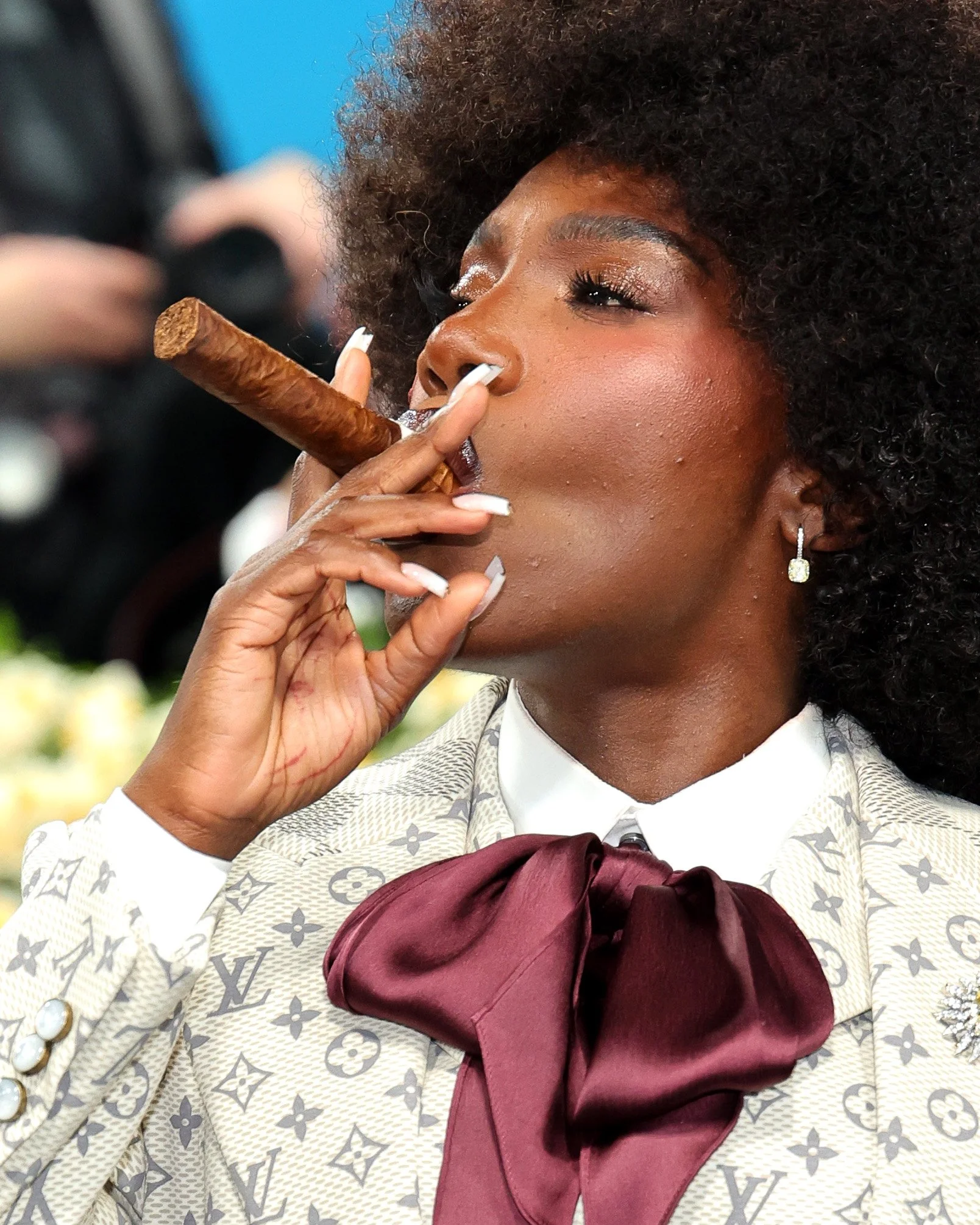 A woman dressed in a designer suit with a Louis Vuitton pattern and a maroon bow, smoking a cigar.