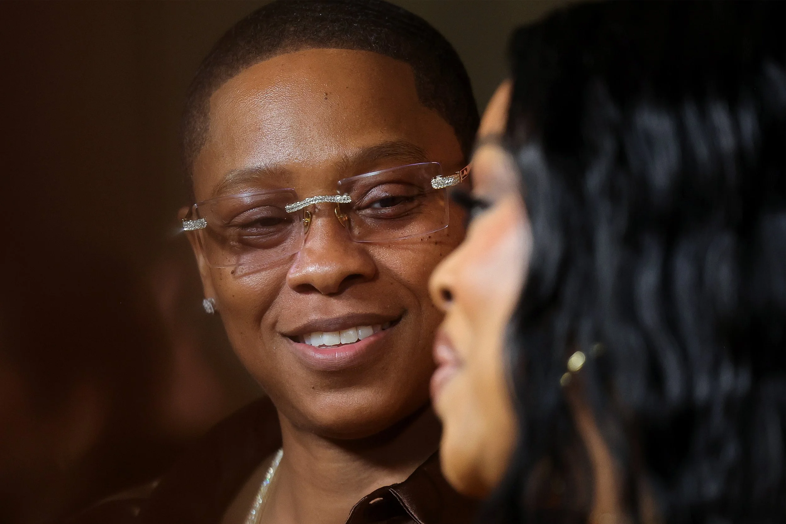 Two women facing each other close up, smiling, one wearing glasses with decorative elements and earrings, the other with dark curly hair and earrings, blurred background