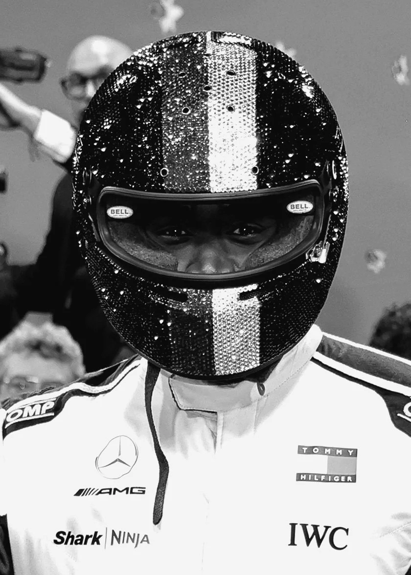 A race car driver wearing a sequined helmet and racing suit with sponsor logos, standing in a garage or pit area.