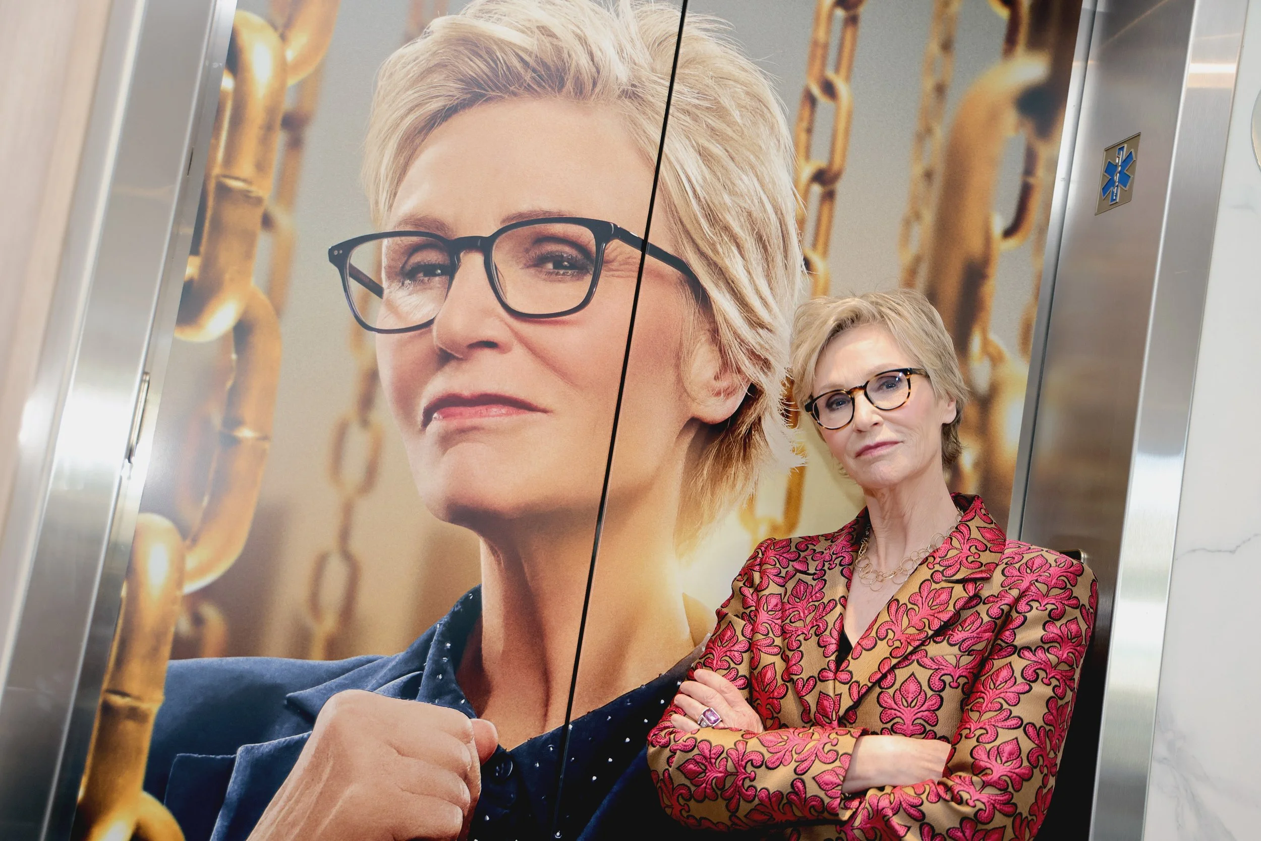 An elderly woman with glasses and blonde hair leaning against an elevator wall with a large poster of herself behind her. The poster features her close-up portrait with glasses, in front of a background of gold chains. The woman is wearing a patterne