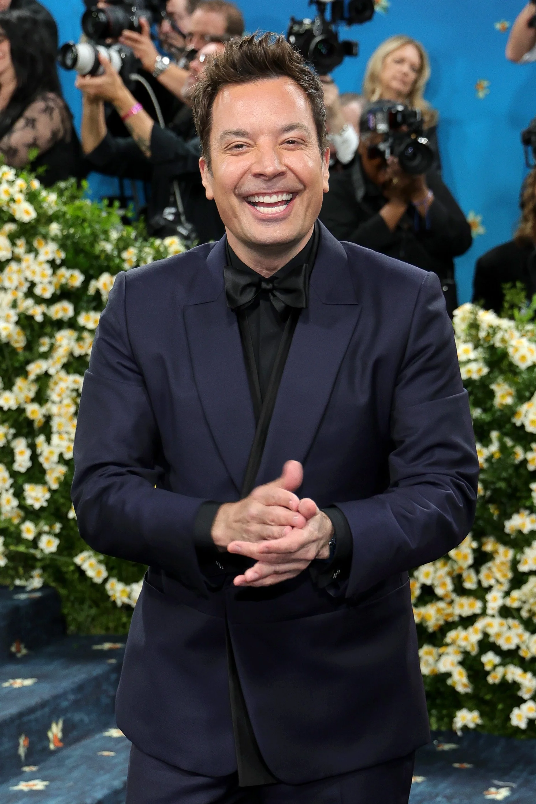 A man in a dark blue tuxedo with a black bow tie, smiling and clapping his hands, at a formal event with photographers and flowers in the background.