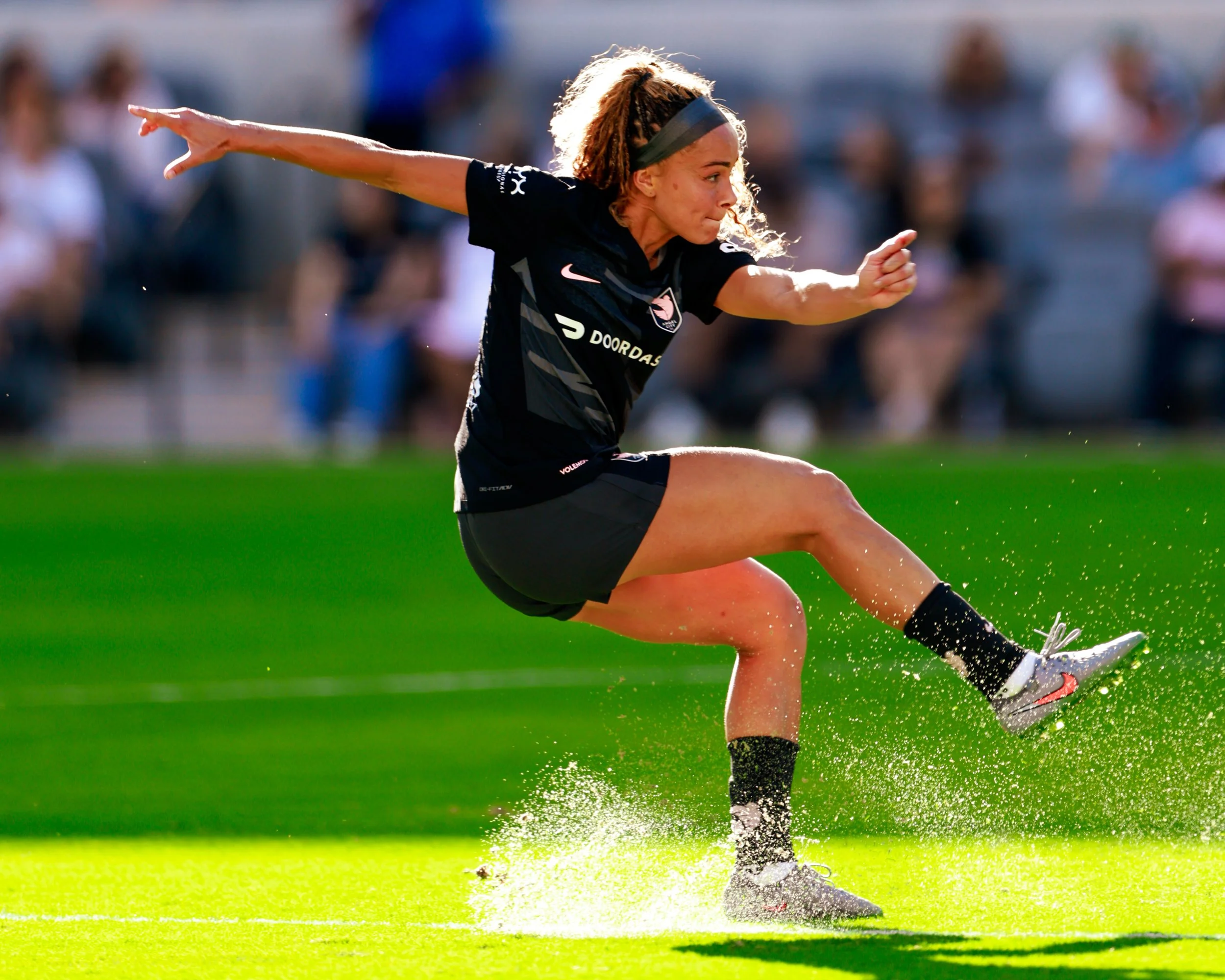 A female soccer player in black uniform kicking a soccer ball on a grass field during a match.
