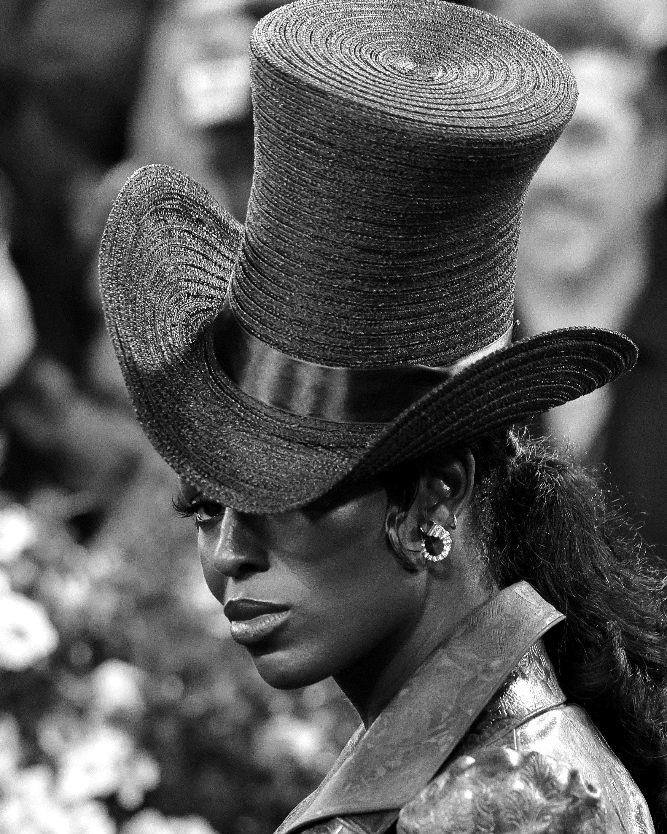 A woman wearing a large, textured, woven hat with a wide brim, and a leather jacket with a patterned texture. She has earrings and is looking over her shoulder, with a blurred background.