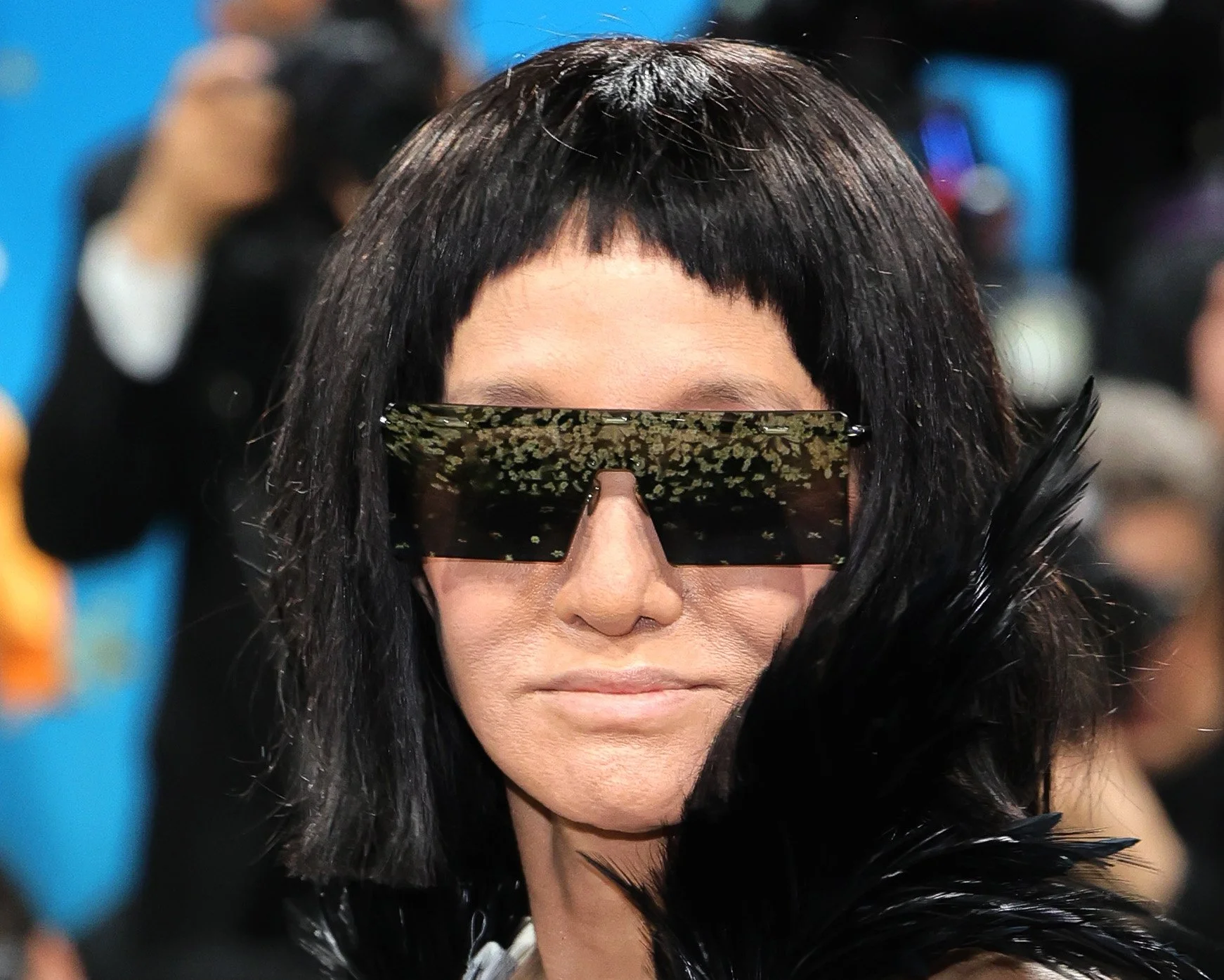 Close-up of a woman with black hair wearing large, futuristic sunglasses with a digital floral pattern, and a feathered accessory on her shoulder.