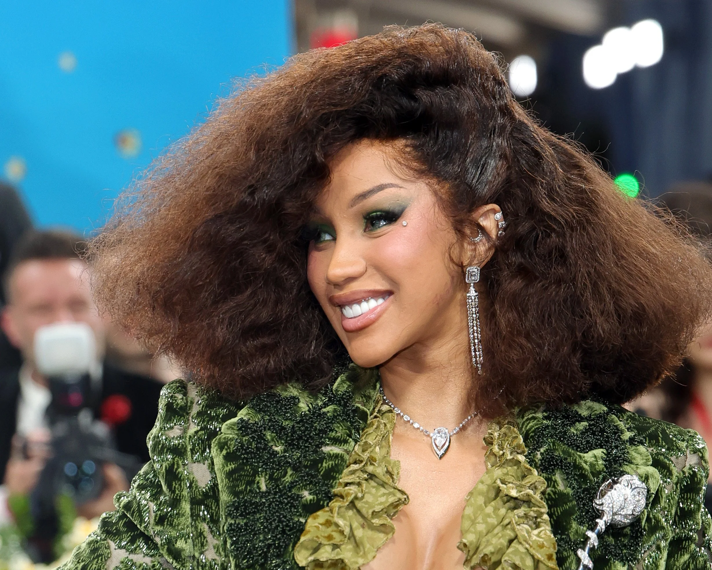 Woman with voluminous curly hair, wearing green eyeshadow and diamond jewelry, smiling at a formal event.