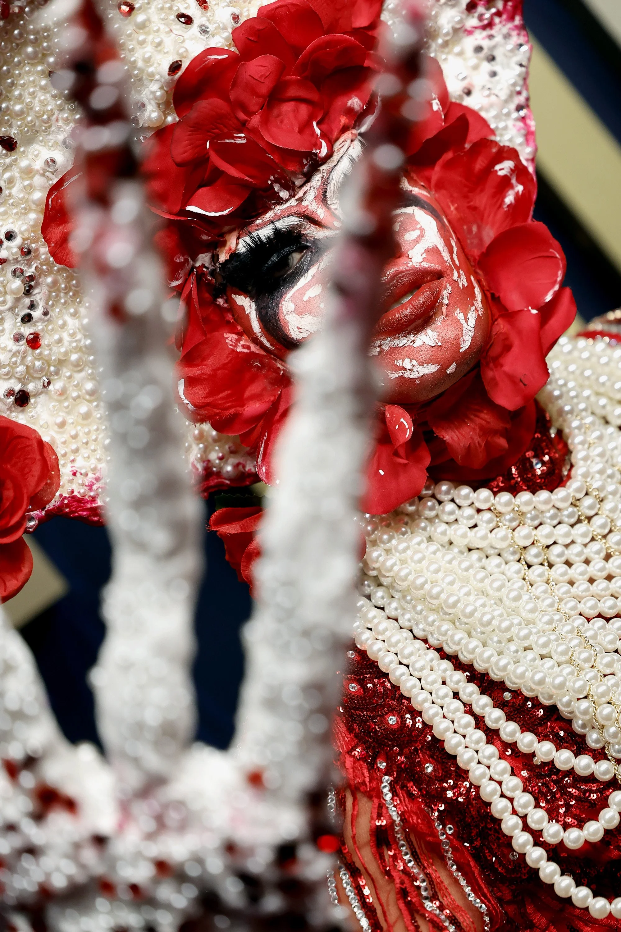 A person in a colorful costume with red petals, pearls, and sequins, wearing clown makeup with exaggerated black eye makeup and red lipstick, behind white decorative bars.