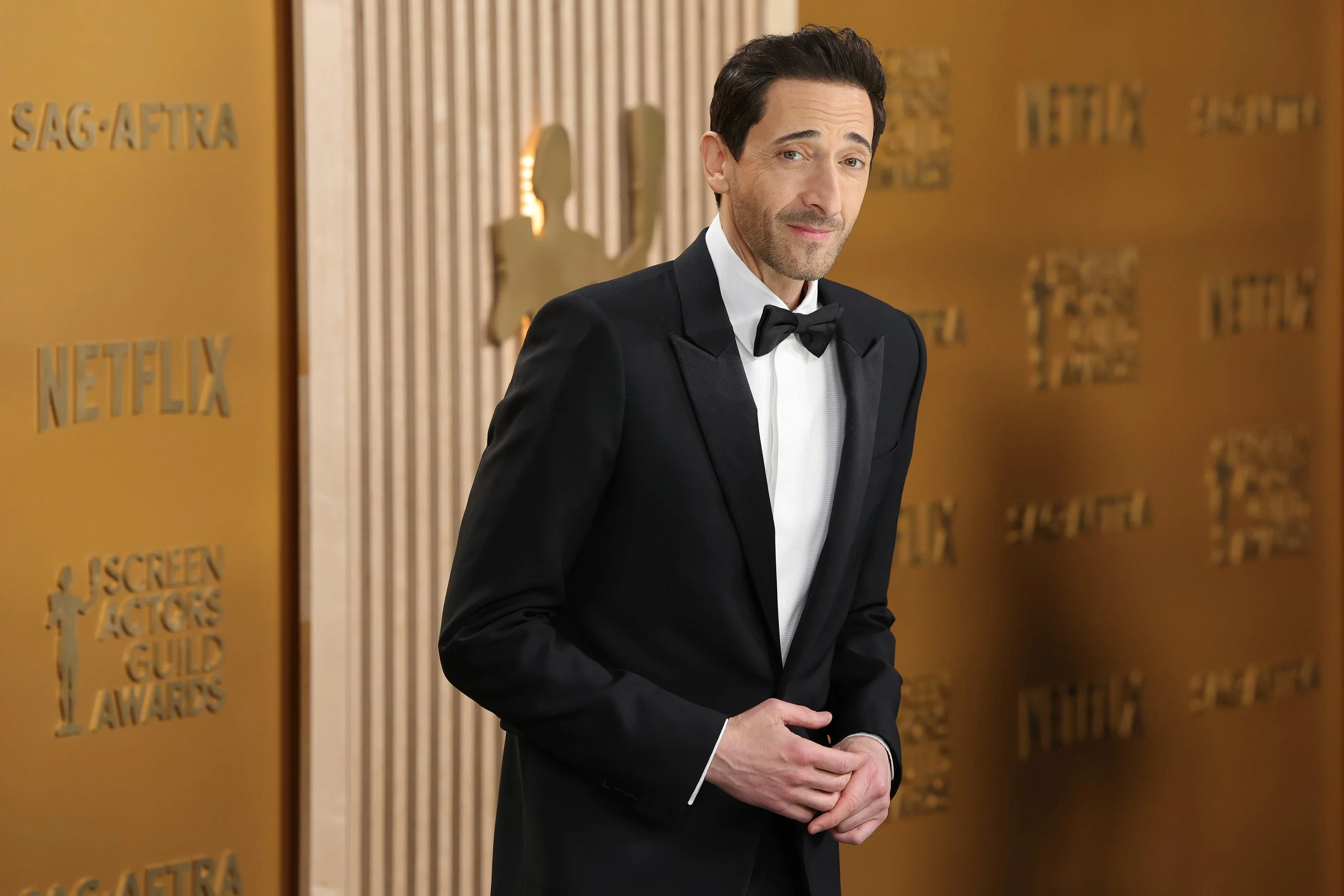 Man in black tuxedo with bow tie at awards event with gold backdrop featuring logos for Netflix and SAG-AFTRA.