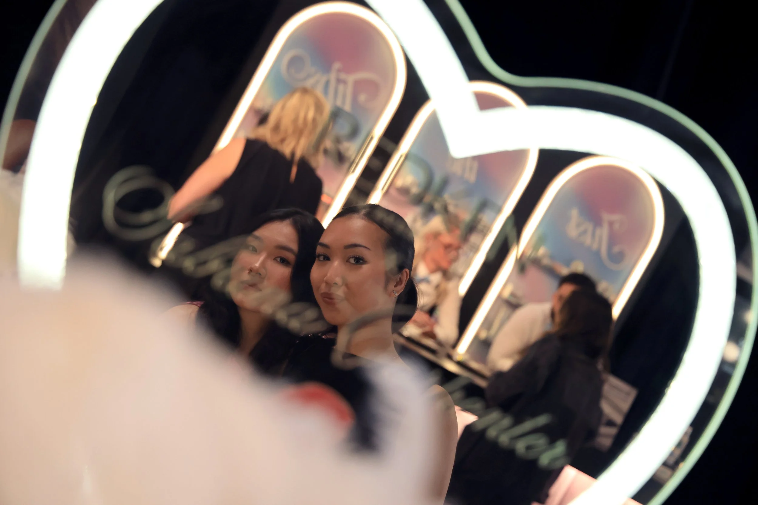 Two women taking a selfie in a mirror at a cafe with a neon sign that says 'Coffee'.